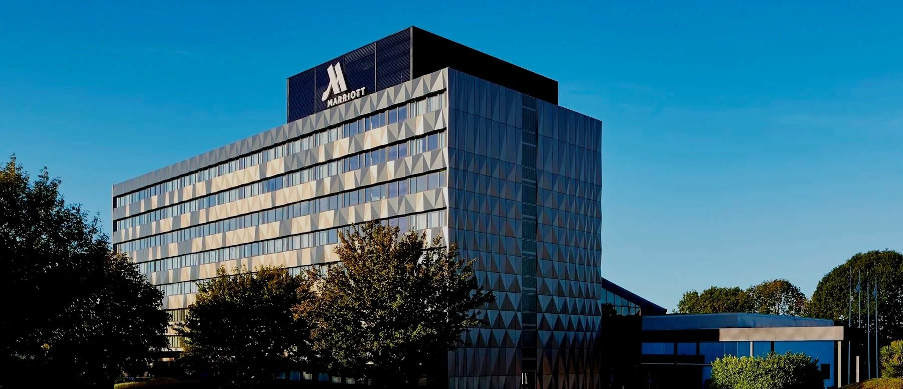 Exterior of a modern Marriott hotel building with a geometric facade, under a clear blue sky, with trees in the foreground.