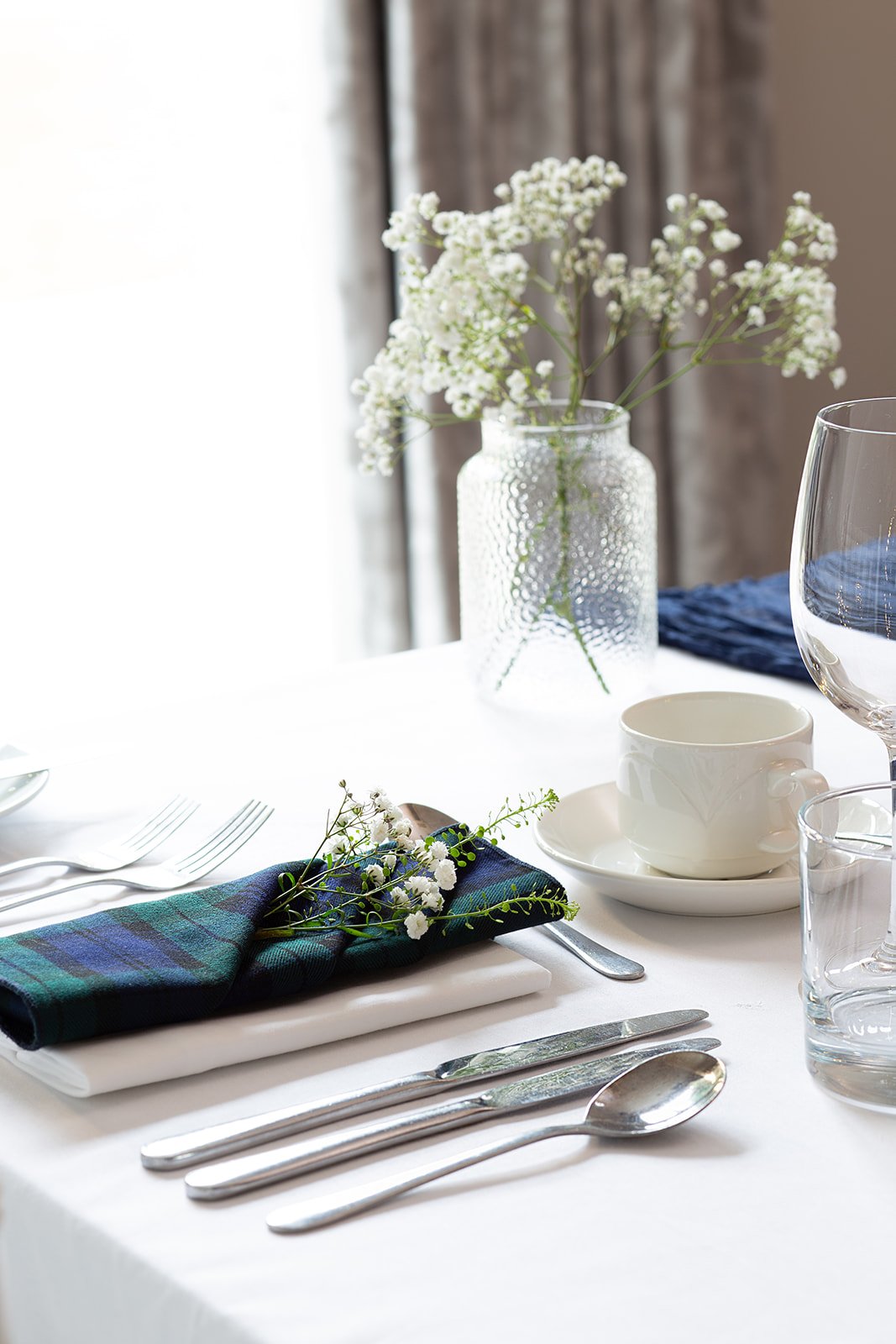 Elegant table setting with a white tablecloth, silverware, a white teacup and saucer, clear drinking glasses, a blue napkin with small white flowers, and a glass vase with white wildflowers in the background.