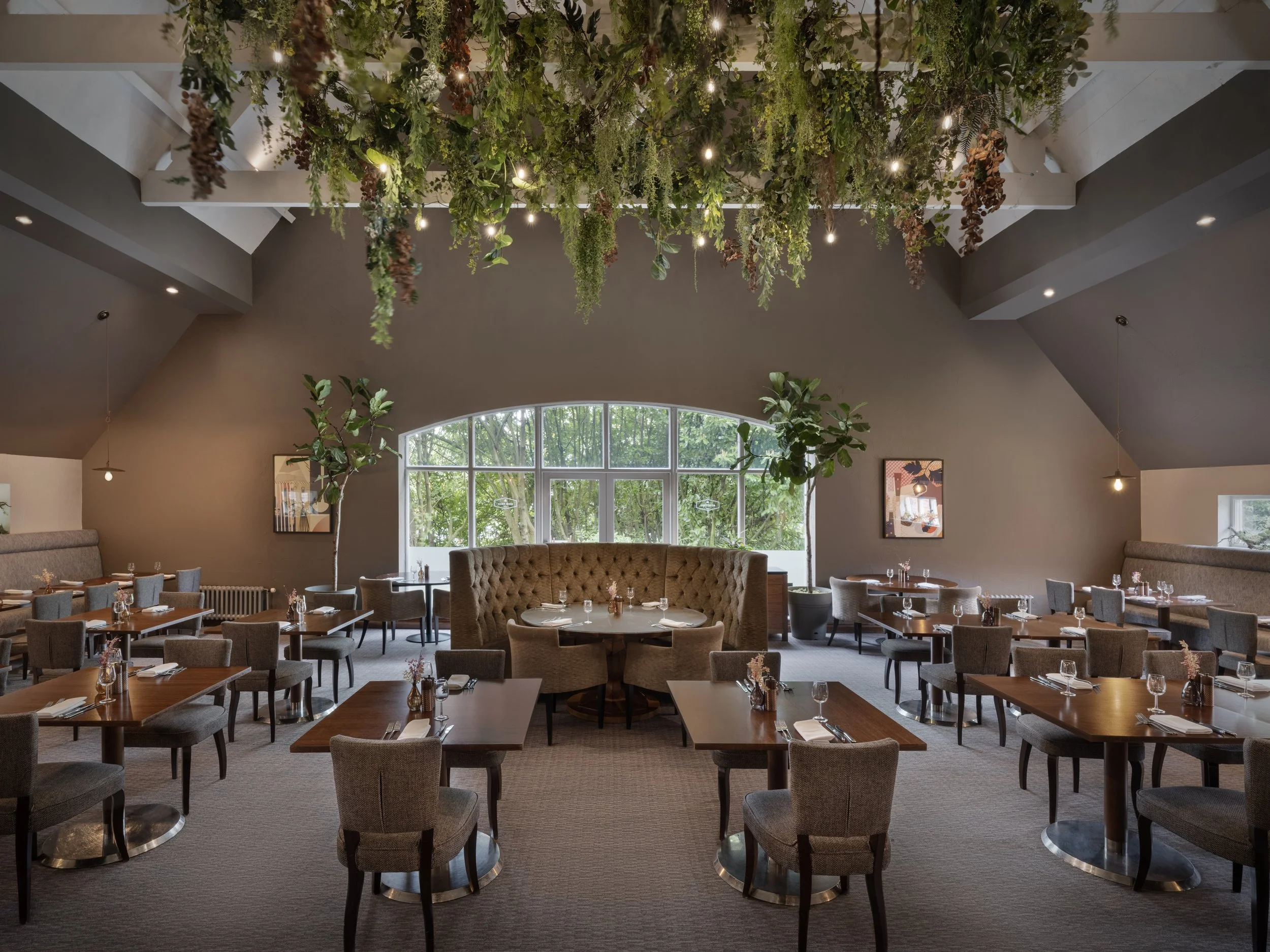 An elegantly decorated restaurant interior featuring a vine and greenery installation on the ceiling with soft lighting, large arched window with trees outside, and neatly set tables with napkins and glassware.