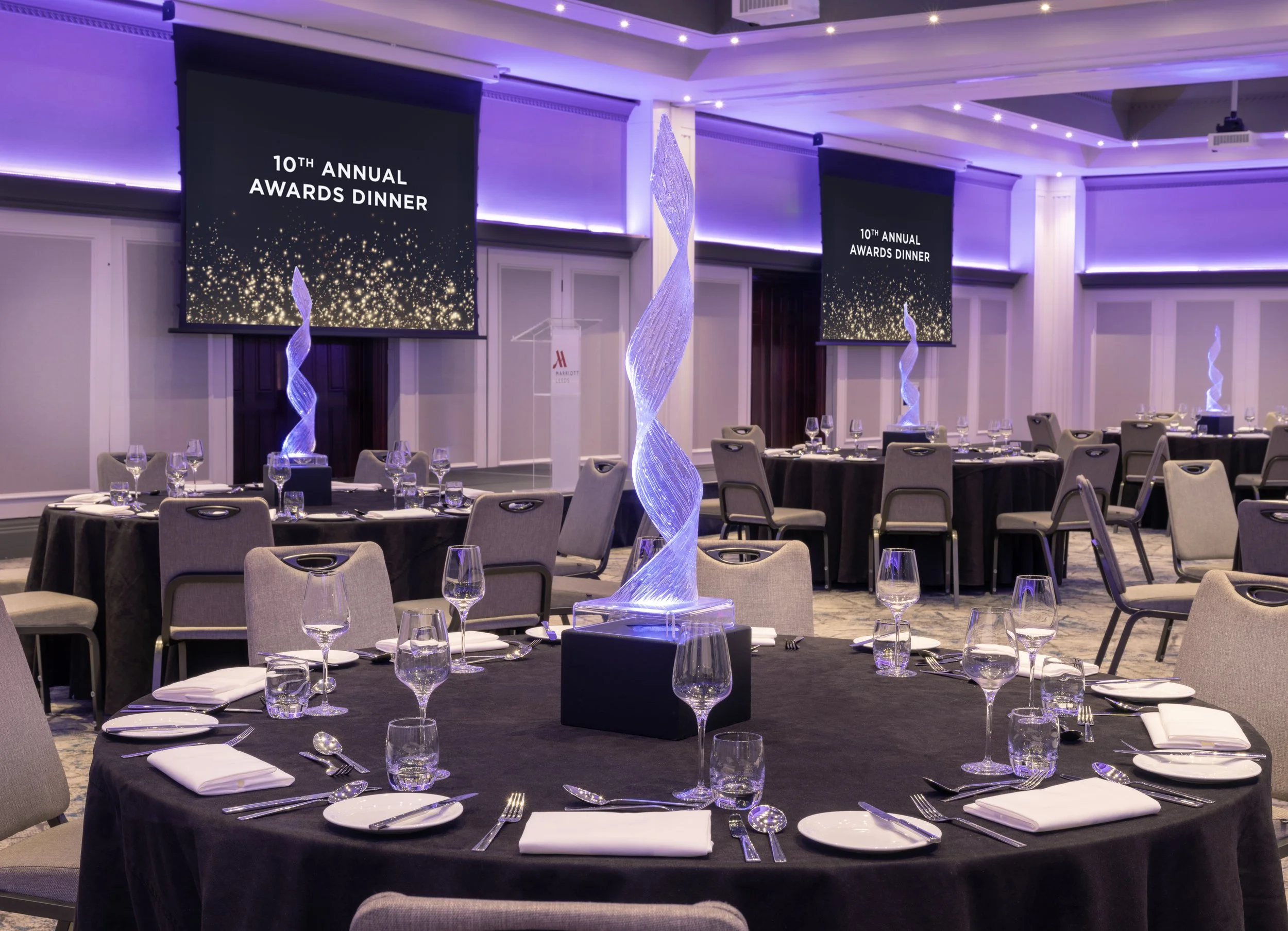Elegant event space set up for a dinner with round tables covered in black tablecloths, each with multiple wine glasses, water glasses, white napkins, and cutlery. The room features tall LED-lit sculptures, large screens displaying '10th Annual Awards Dinner,' purple ambient lighting, and a clear podium in the background.