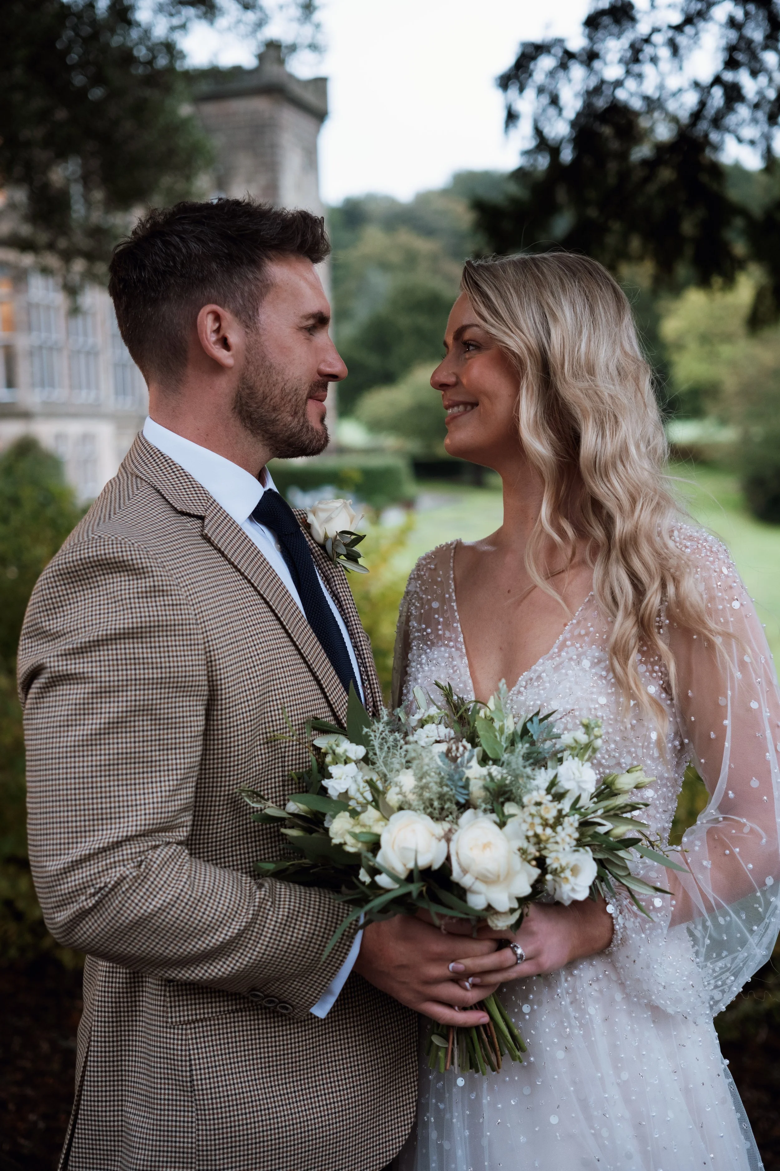 A bride and groom standing close together outdoors, gazing into each other's eyes, with the bride holding a large bouquet of white flowers, wearing a wedding dress, and the groom in a checked blazer, in a park-like setting with historic building in the background.