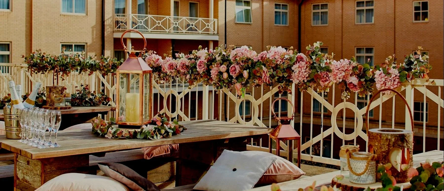 Decorated outdoor balcony with pink flowers, candles, lanterns, glasses, and pillows for a celebration or gathering.