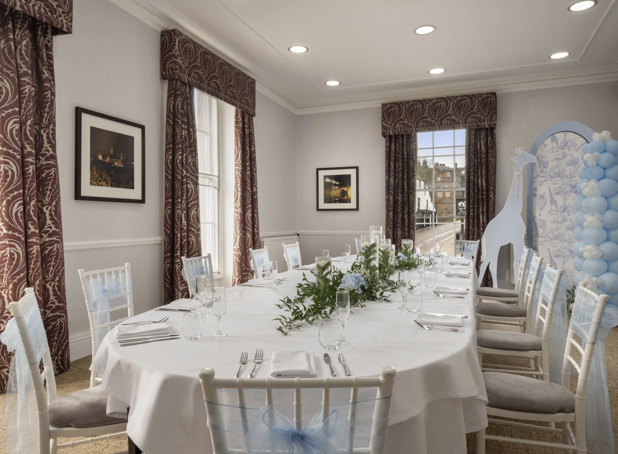 A dining room set up for a celebration with a long table covered with a white tablecloth, decorated with greenery, glassware, and silverware. White chairs with padded seats surround the table. The room has large windows with patterned maroon curtains, framed pictures on the walls, and recessed ceiling lights. A decorative element with blue and white balloons and giraffe illustrations is partially visible on the right.