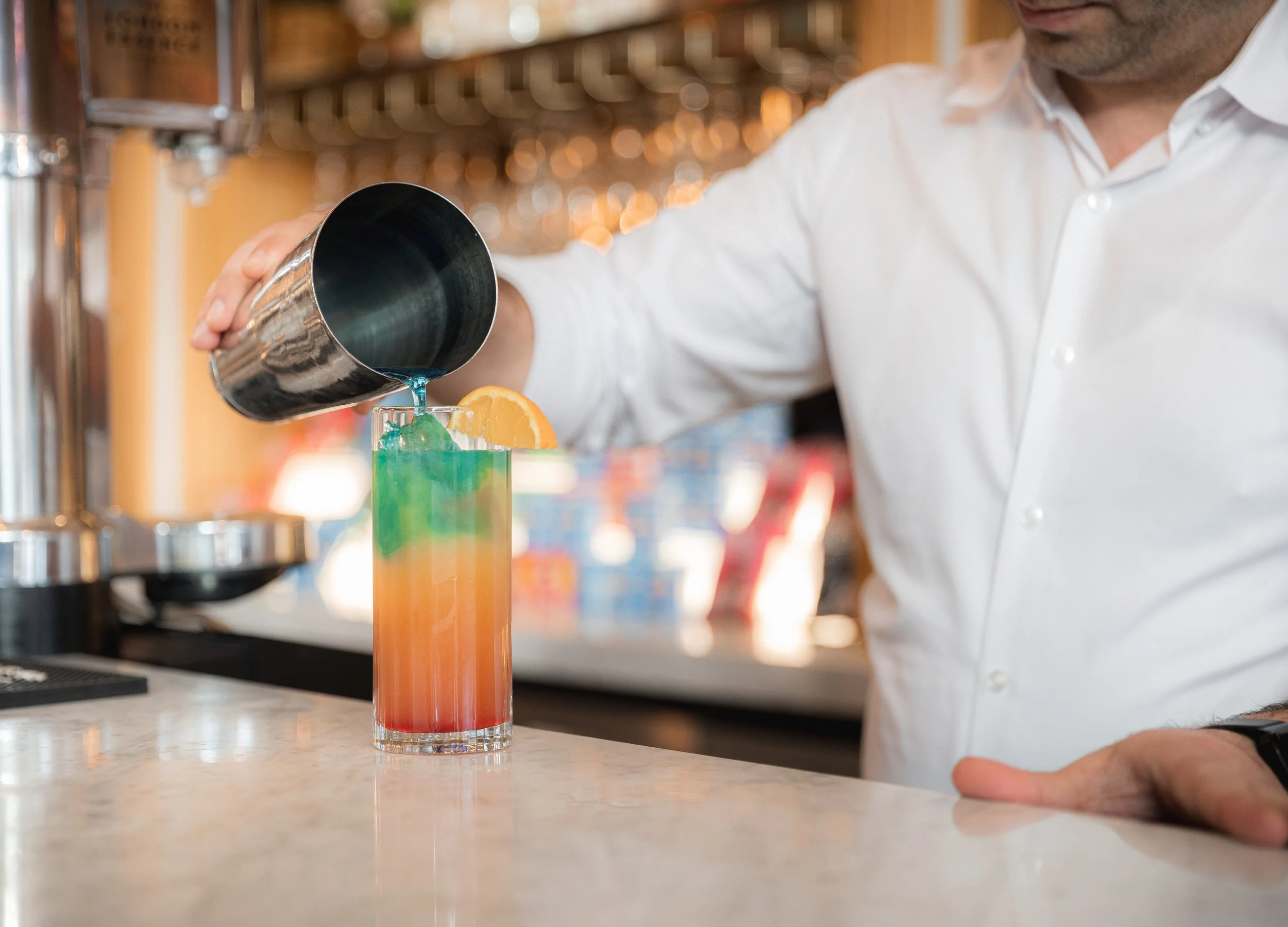 Bartender pouring a blue cocktail from a shaker into a tall glass with an orange slice garnish.