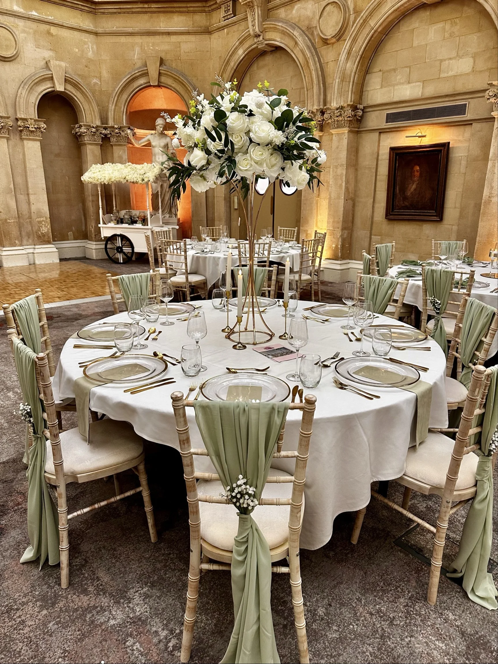 Elegant round banquet table decorated with a tall floral arrangement of white roses and greenery, set with gold-rimmed plates, glassware, and gold flatware, in a historic stone hall with arched doorways, classical sculptures, and framed portrait.
