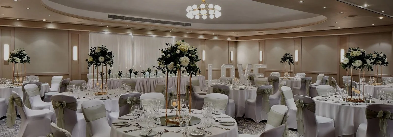 Elegant wedding reception hall decorated with tall floral centerpieces and a large love sign in the background.