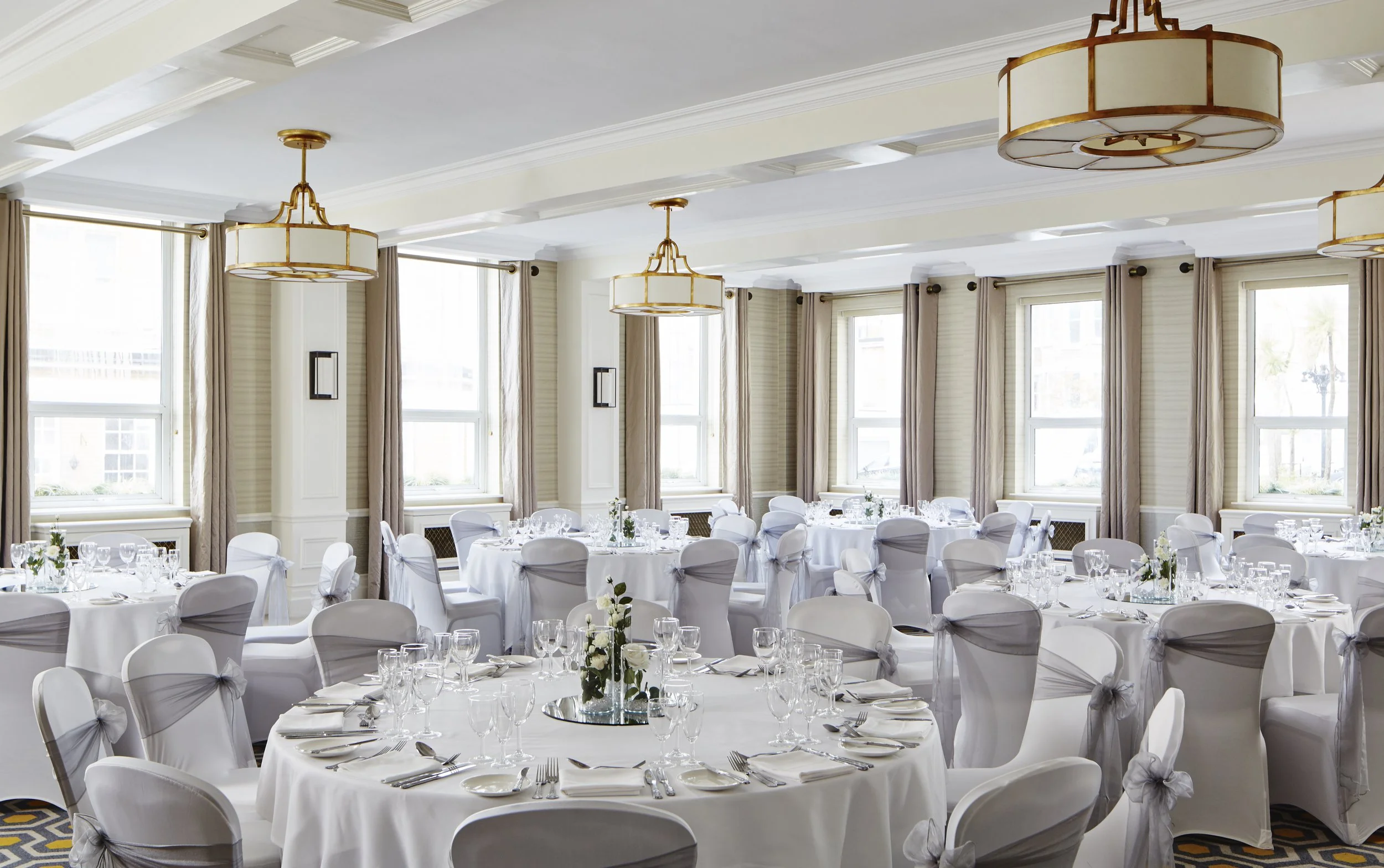 Elegant banquet hall with round tables covered in white tablecloths, set with glassware and silverware, and decorated with floral centerpieces. The room features large windows with cream curtains, white walls, crown molding, and modern hanging light fixtures.