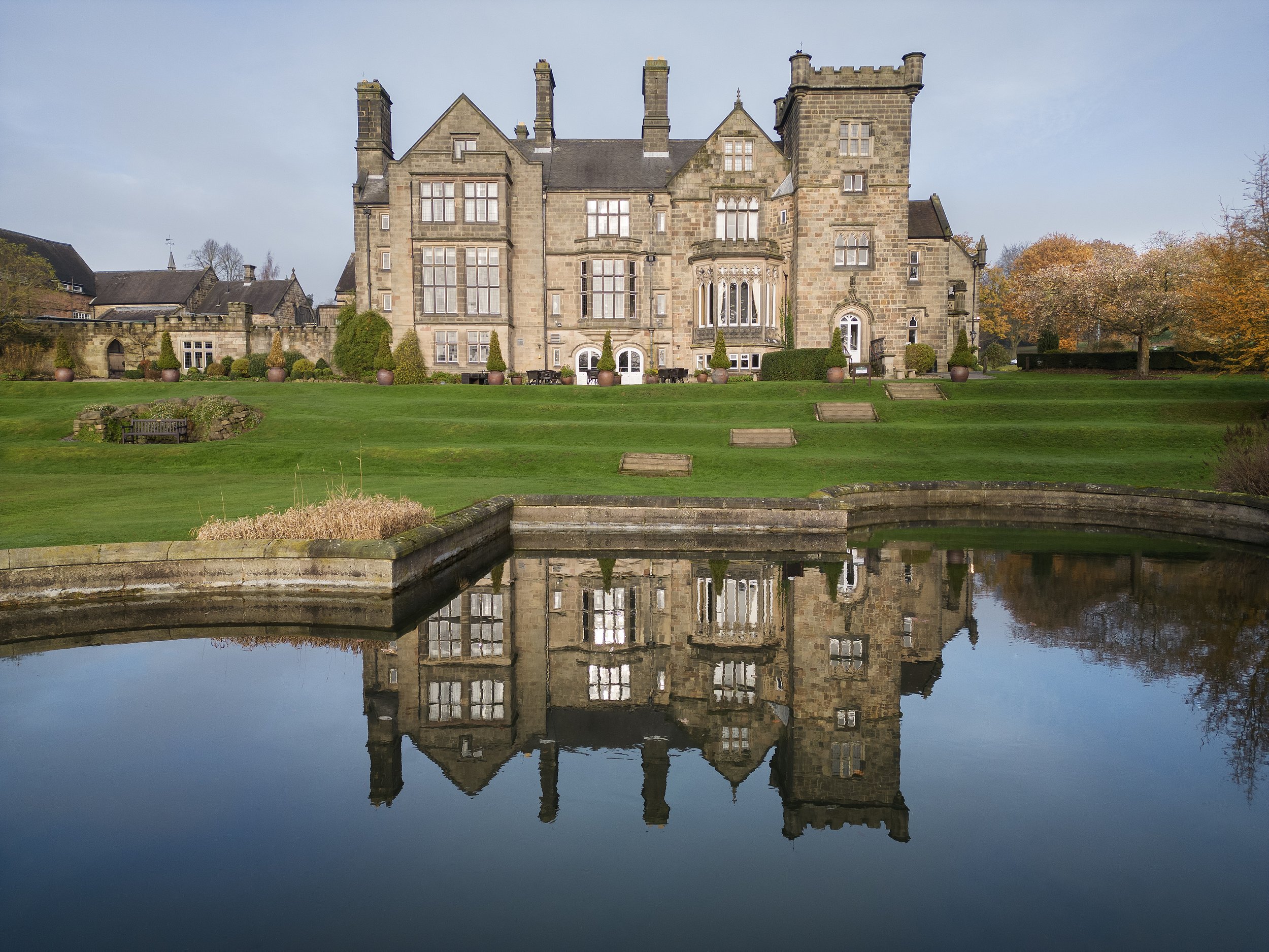 A large stone mansion reflects in a calm body of water surrounded by a well-manicured lawn and autumn-colored trees.