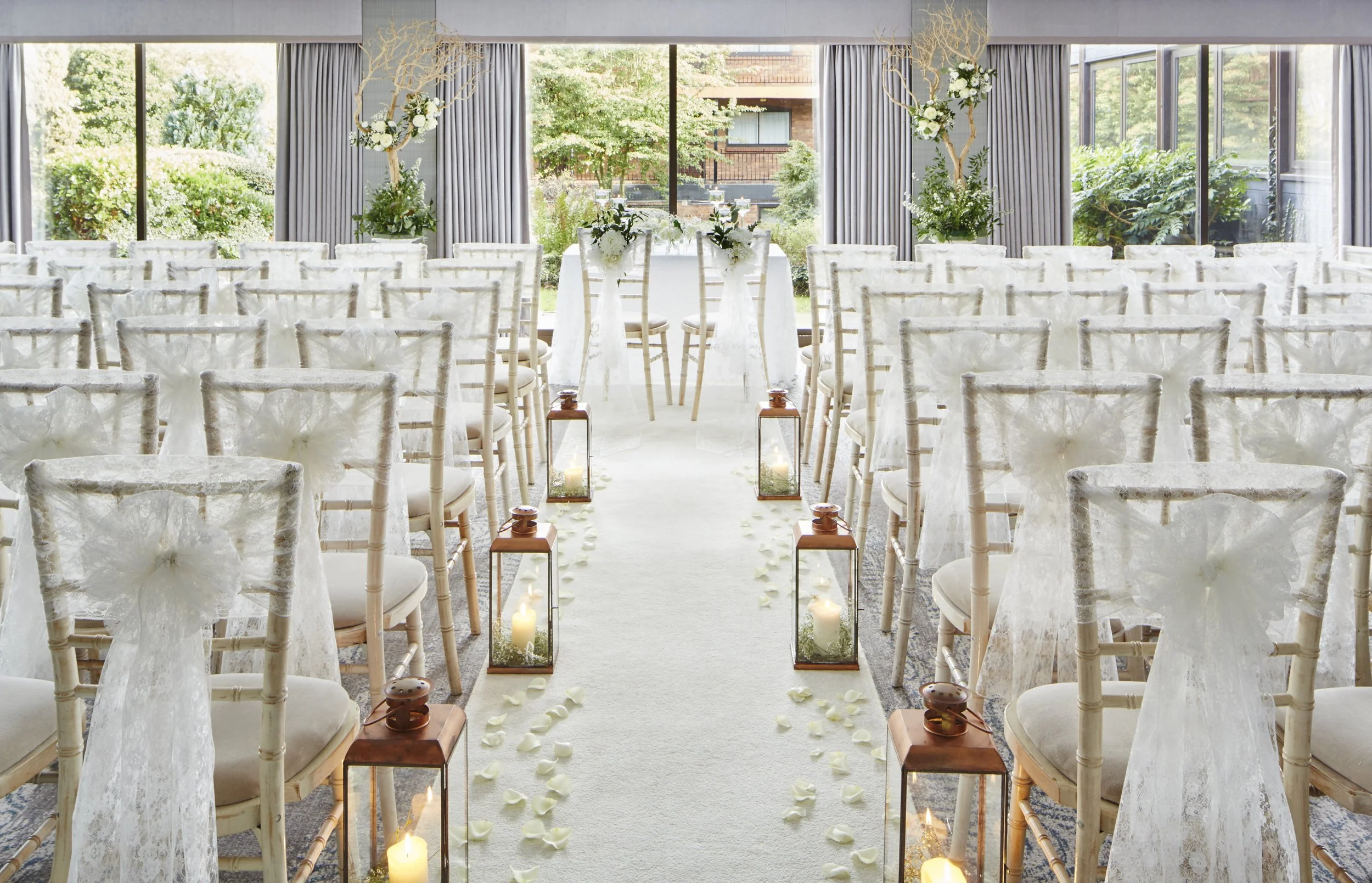 Interior of a wedding ceremony setup with rows of white covered chairs facing an aisle decorated with candles and flower petals, large windows with outdoor greenery in the background.