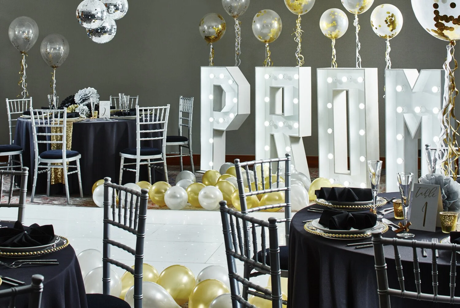 Elegant party or celebration setup with black and gold table settings, black napkins, gold and black decor, balloons, and large illuminated letters spelling 'PROM' in the background.