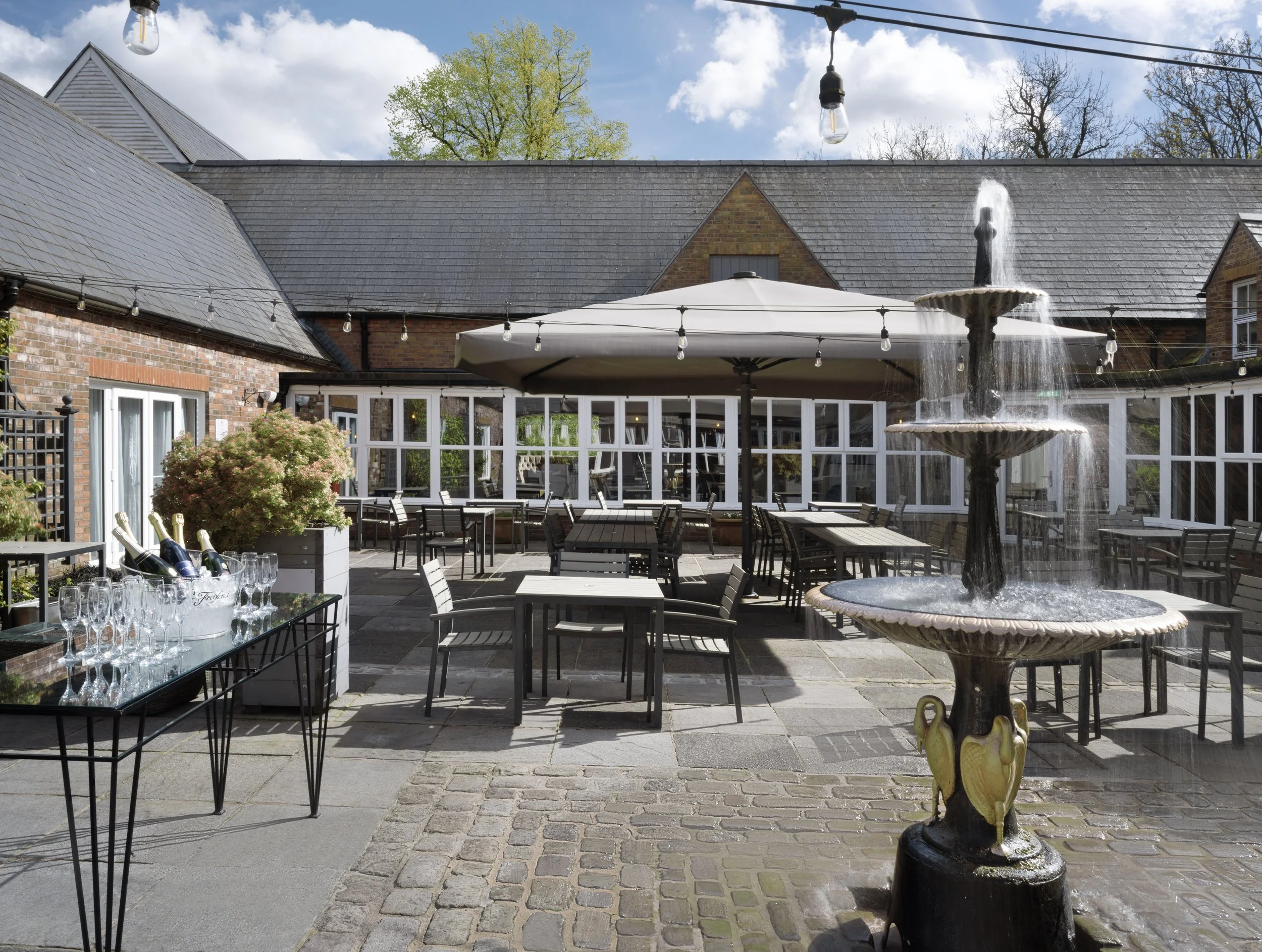 An outdoor patio area with a fountain in the foreground, surrounded by tables and chairs. String lights are hanging above, and there is a large umbrella providing shade. The background features brick buildings with pitched roofs and trees under a partly cloudy sky.