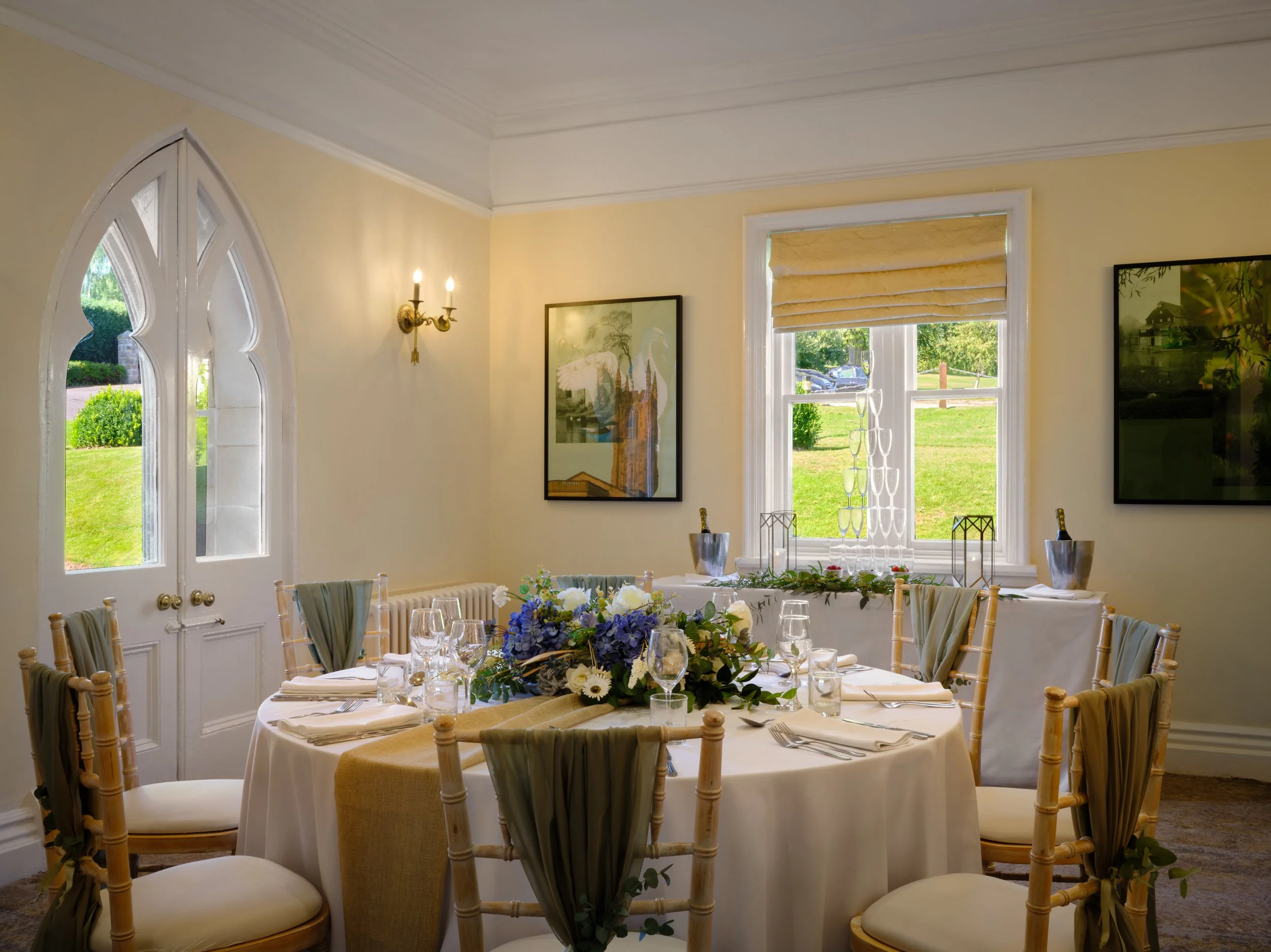 An elegant room decorated for a formal event, featuring a round table with a floral centerpiece, surrounded by wooden chairs with white cushions, large windows with beige Roman shades, and framed artwork on the walls.