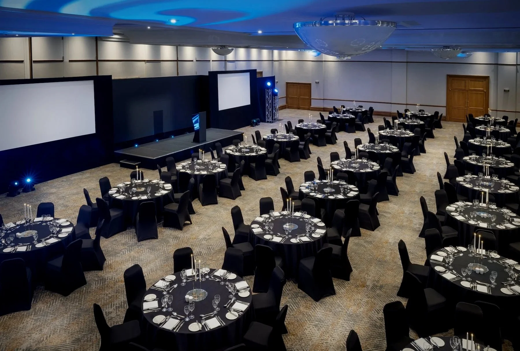 Large banquet hall with round tables covered in black tablecloths, set with fine dinnerware, glasses, and candles. The stage has large screens and lighting equipment, indicating setup for a formal event or conference. The room has chandeliers and wood-paneled doors.