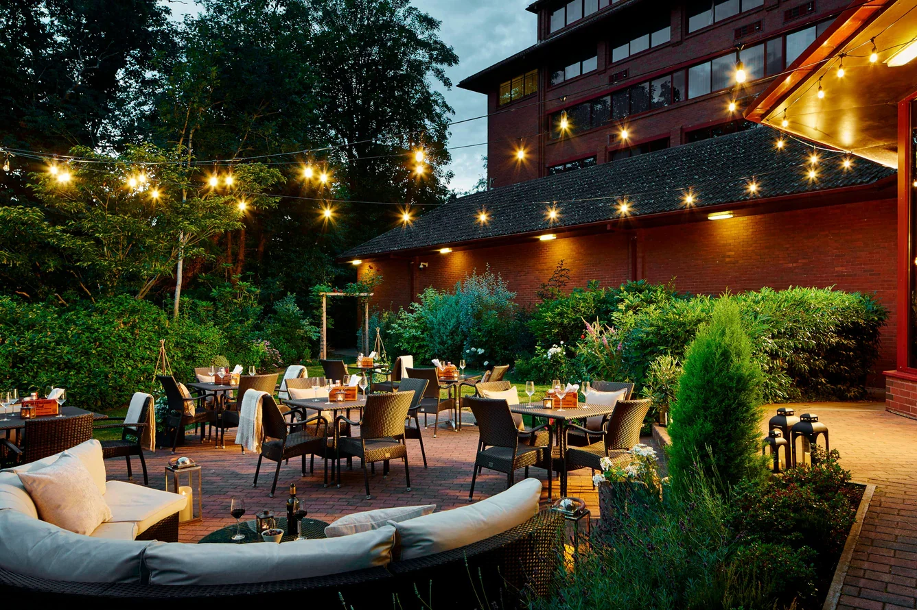 Outdoor dining area with tables and chairs, string lights overhead, surrounded by lush greenery and a brick building in the evening.