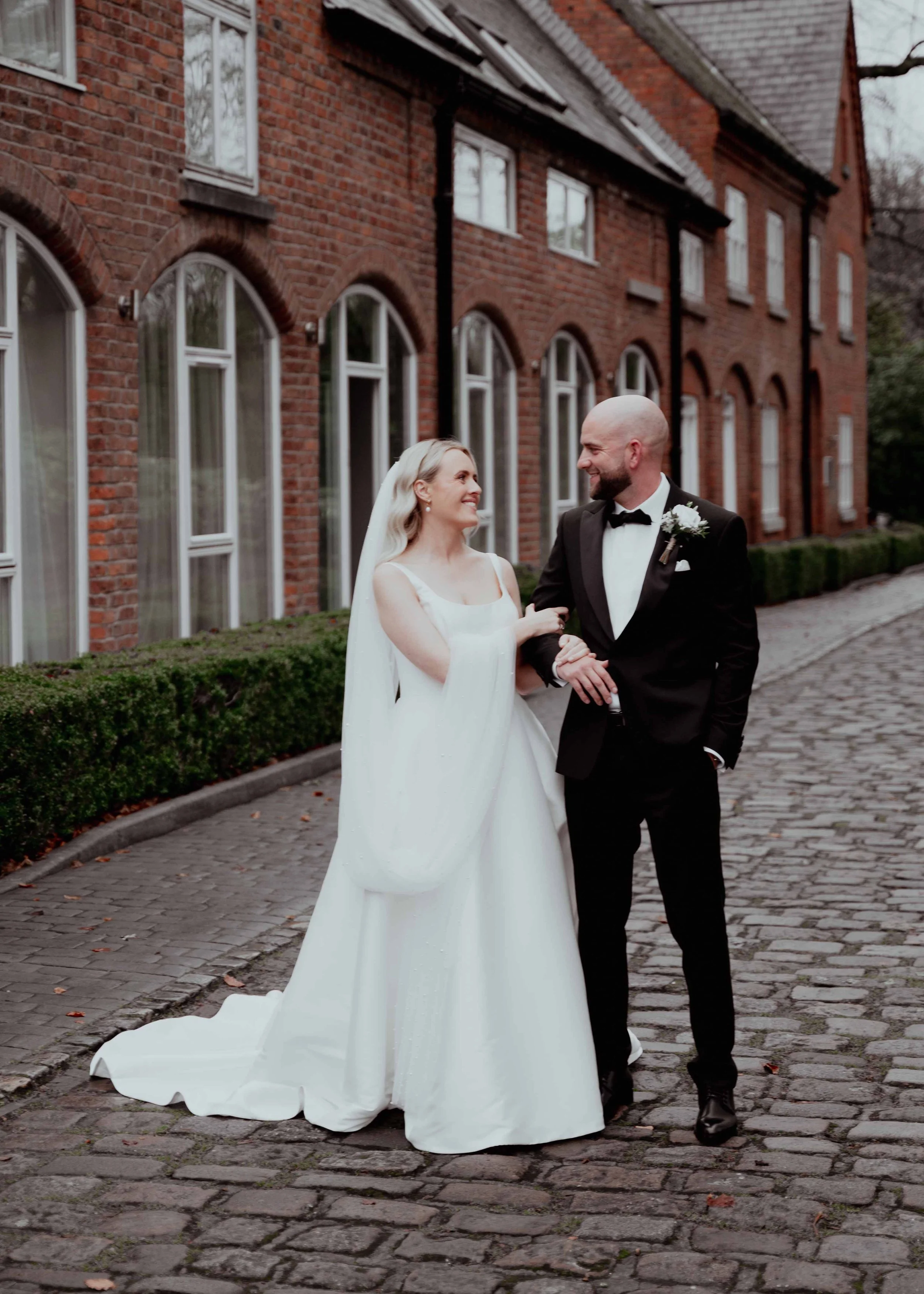 A bride and groom smile at each other while walking on a cobblestone street in front of a red brick building with large arched windows. The bride is wearing a white wedding dress and veil, and the groom is dressed in a black tuxedo with a bow tie and boutonniere.