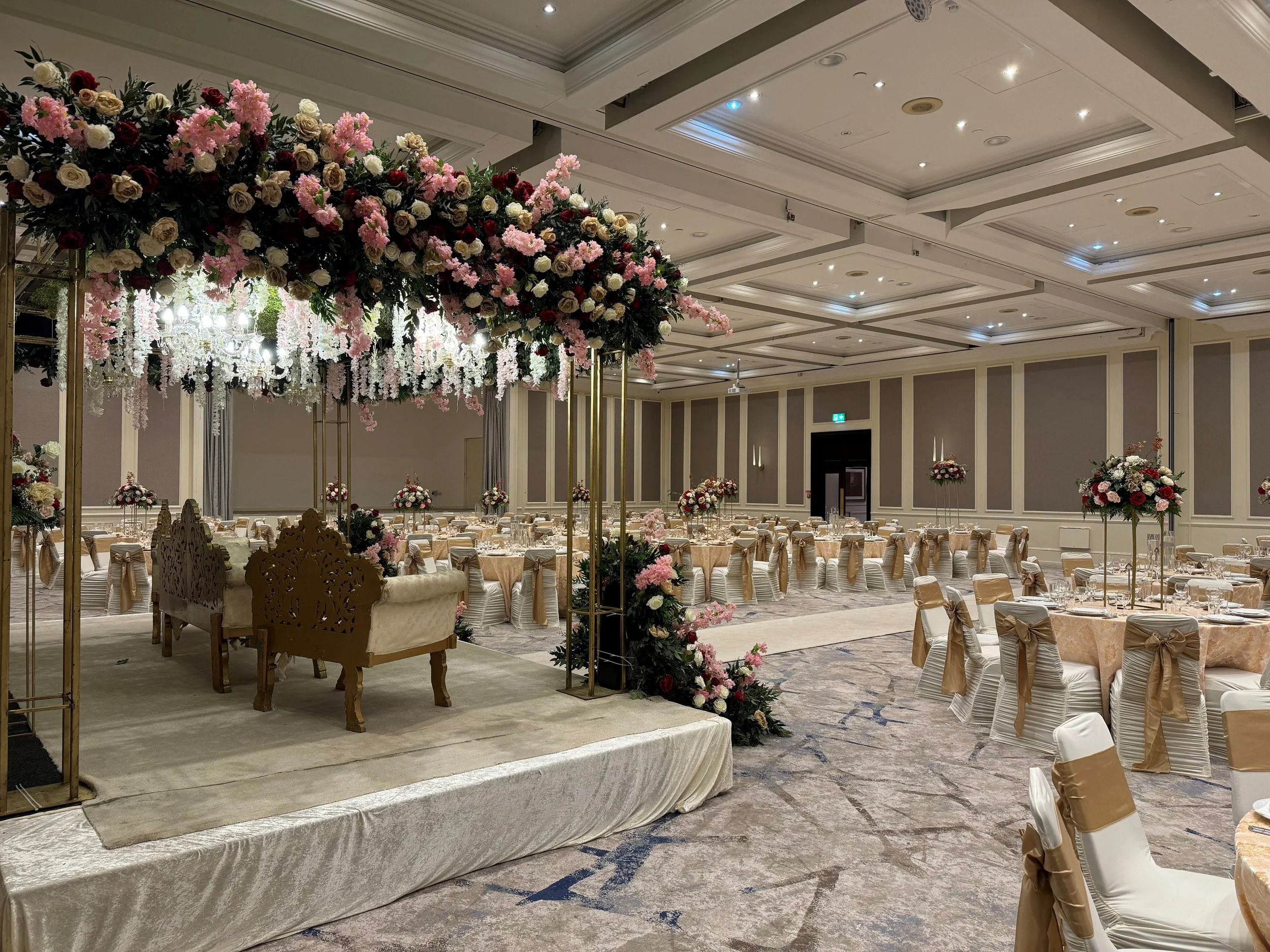 An elegant banquet hall decorated for a wedding reception with round tables covered in beige tablecloths and chairs adorned with beige bows. A large floral centerpiece with pink, cream, and red flowers is suspended above the main stage area, which features ornate gold chairs and a beige carpet.