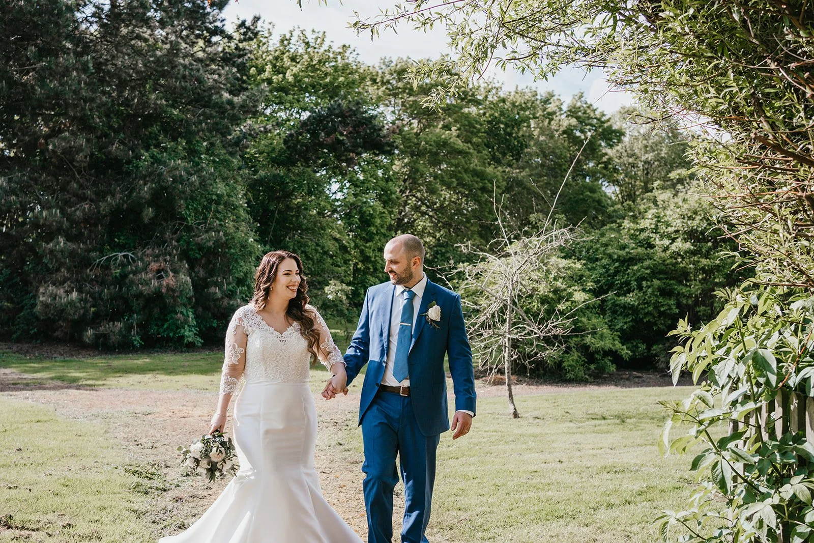 A bride and groom walking hand in hand outdoors on a grassy area, smiling at each other with trees in the background during a wedding celebration.