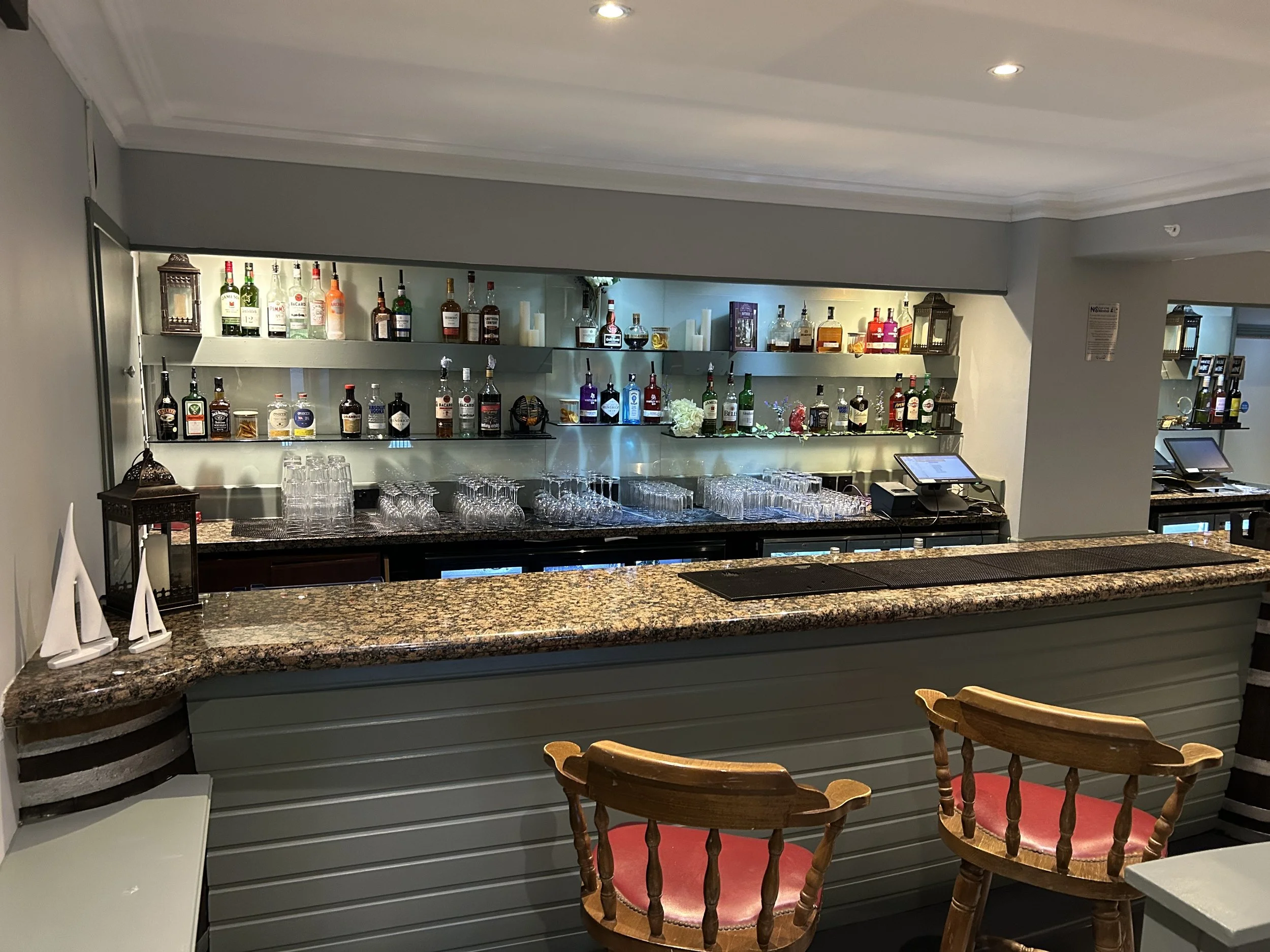 A bar area with a granite countertop, glasses, and shelves stocked with various bottles of alcohol. There are wooden chairs with red cushions in front of the bar.