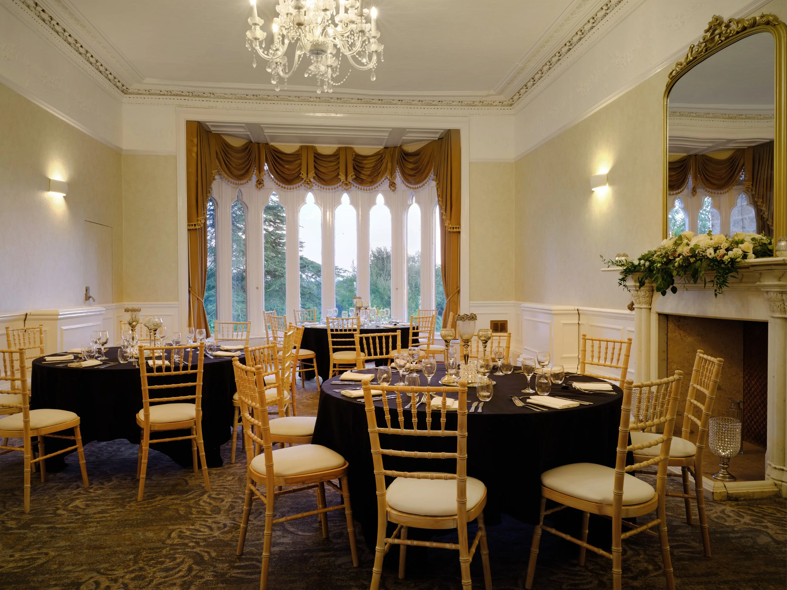 Elegant banquet hall decorated for a special event with round tables covered in black tablecloths, gold chairs, and table settings. Large windows with ornate gold curtains and a chandelier hanging from the decorated ceiling.