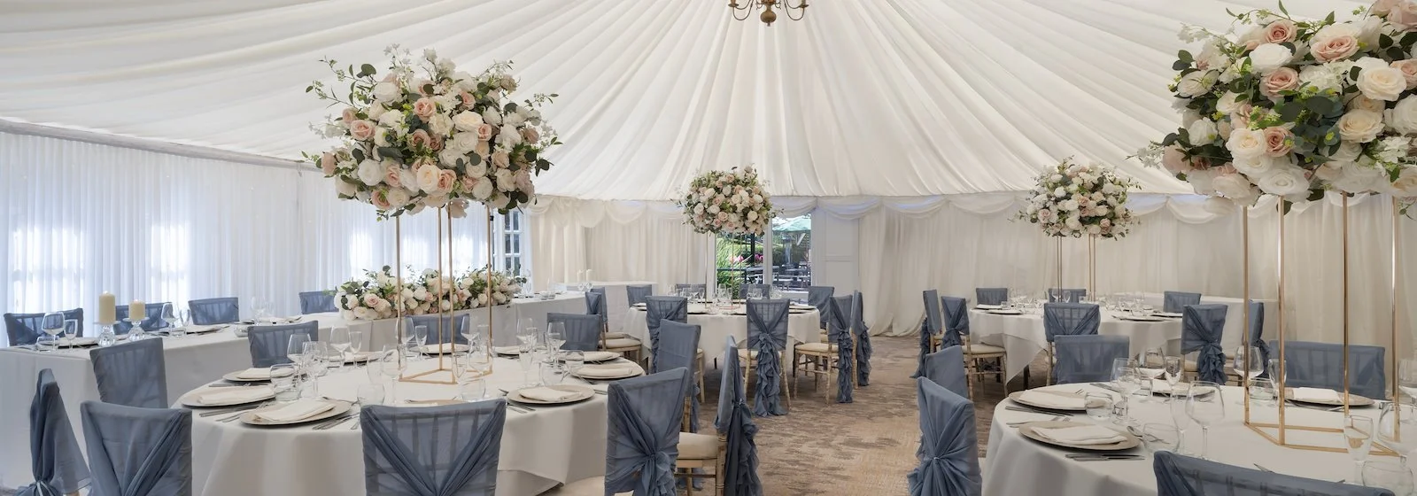Elegant wedding reception tent decorated with large floral centerpieces on tall stands, round tables with white tablecloths, and chairs with blue fabric covers tied at the back. Soft natural light filters through sheer white drapes.
