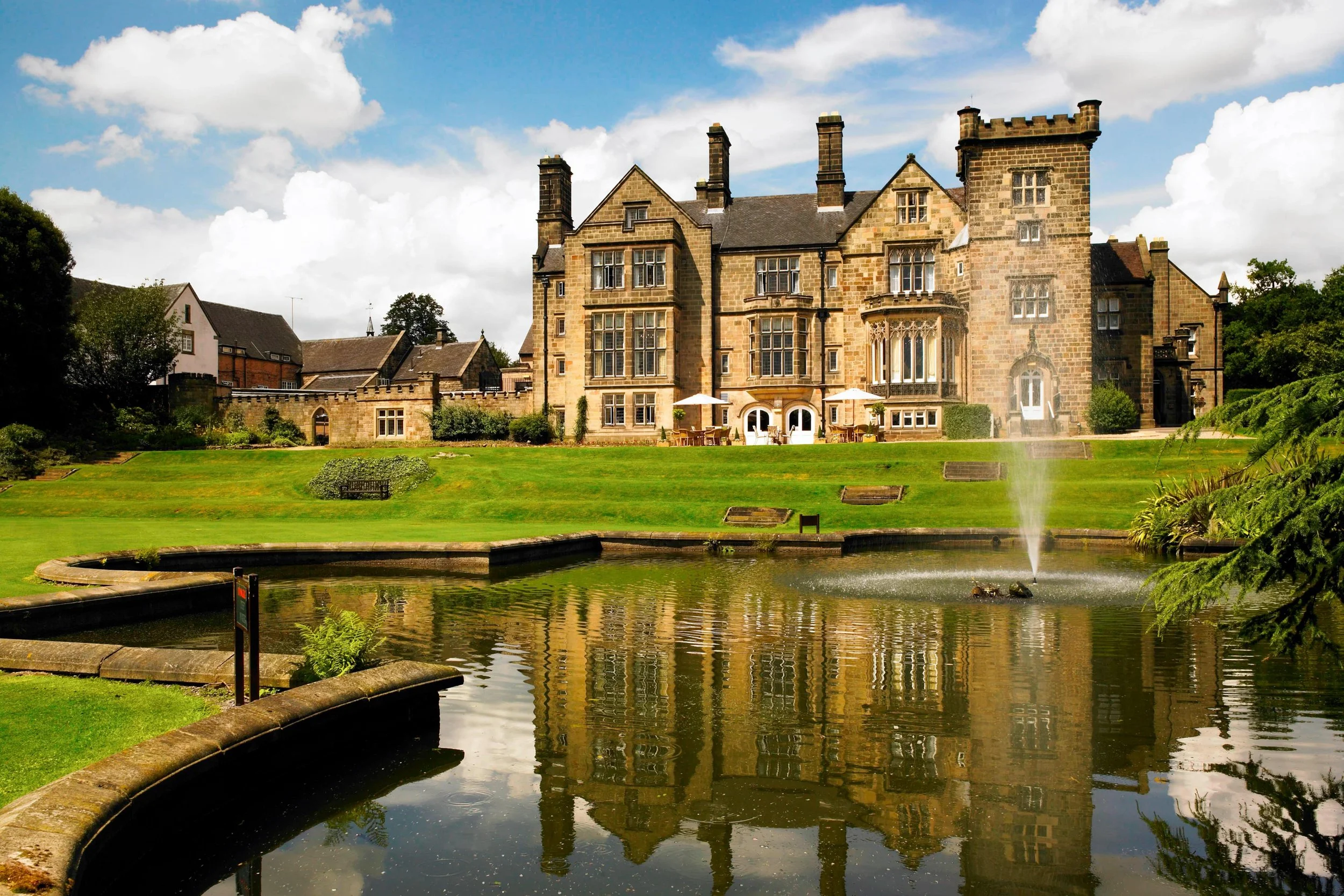 A large historic stone mansion with multiple tall chimneys, surrounded by a well-maintained lawn and garden. In the foreground, a fountain in a pond reflects the house and the partly cloudy sky.