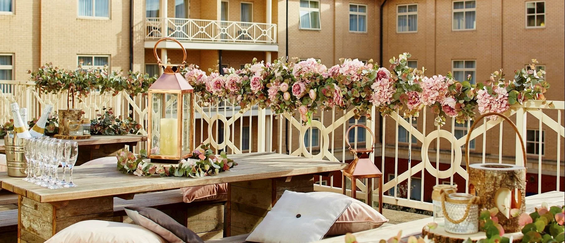 Decorated outdoor balcony with pink floral garland, lantern, champagne glasses, and pillows.
