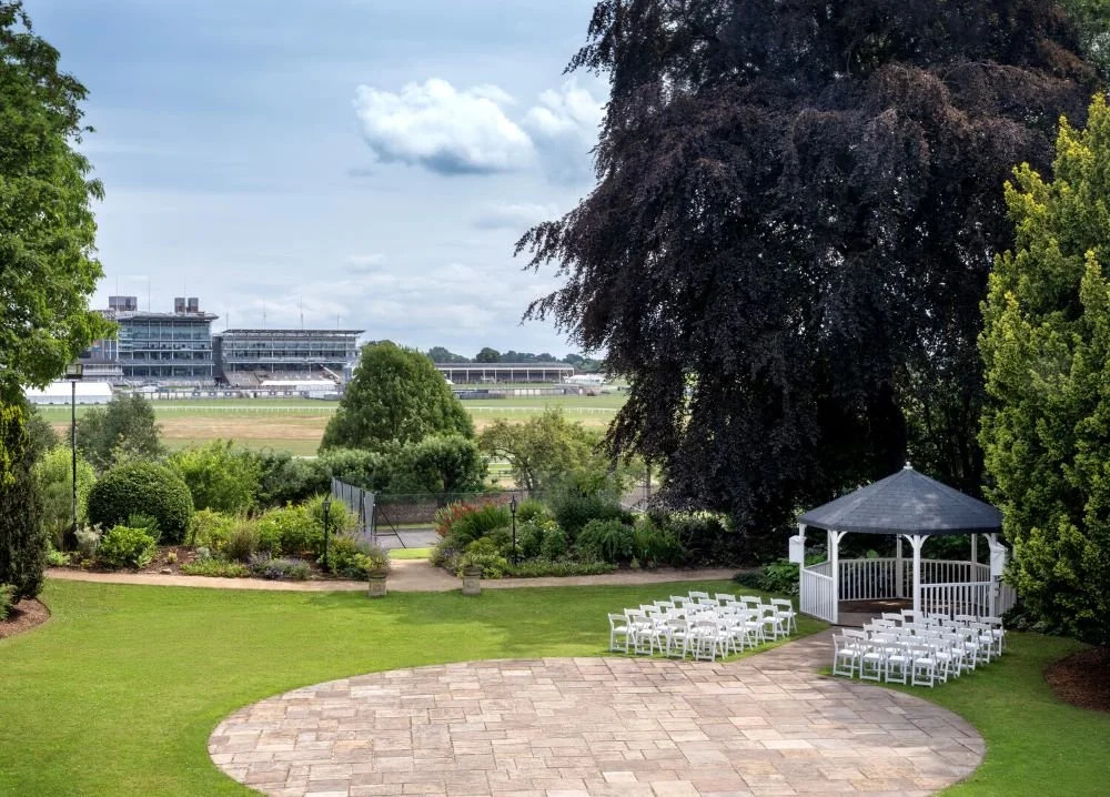 A garden with a gazebo and white chairs, overlooking a racetrack and grandstand in the distance.
