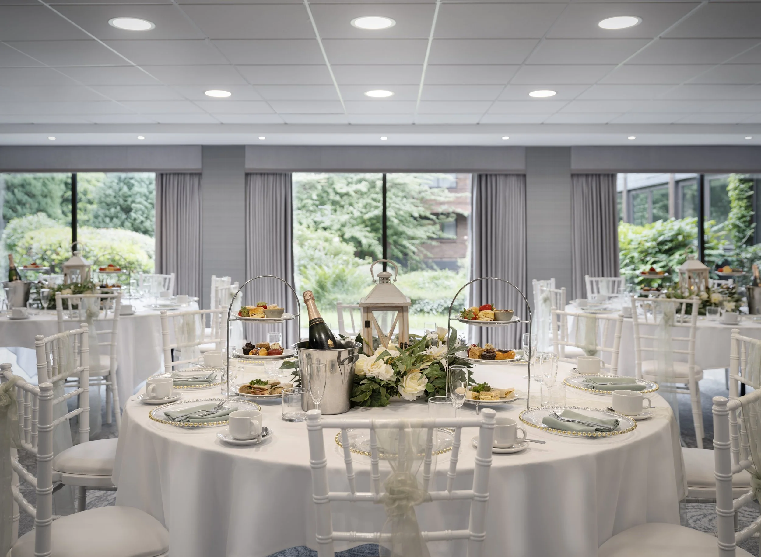 Elegant banquet table setup with white china, silverware, and floral centerpiece, set in a bright room with large windows and gray curtains.