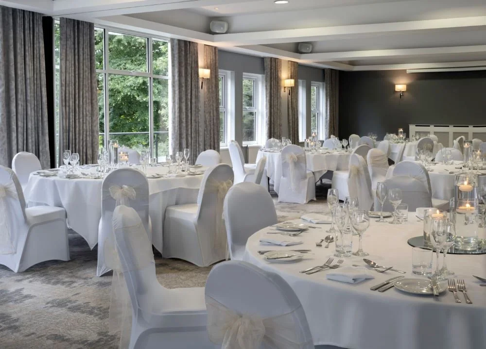 Elegant banquet hall decorated for a formal event with round tables covered in white tablecloths, white chairs tied with bows, and candle centerpieces, with large windows and gray curtains.