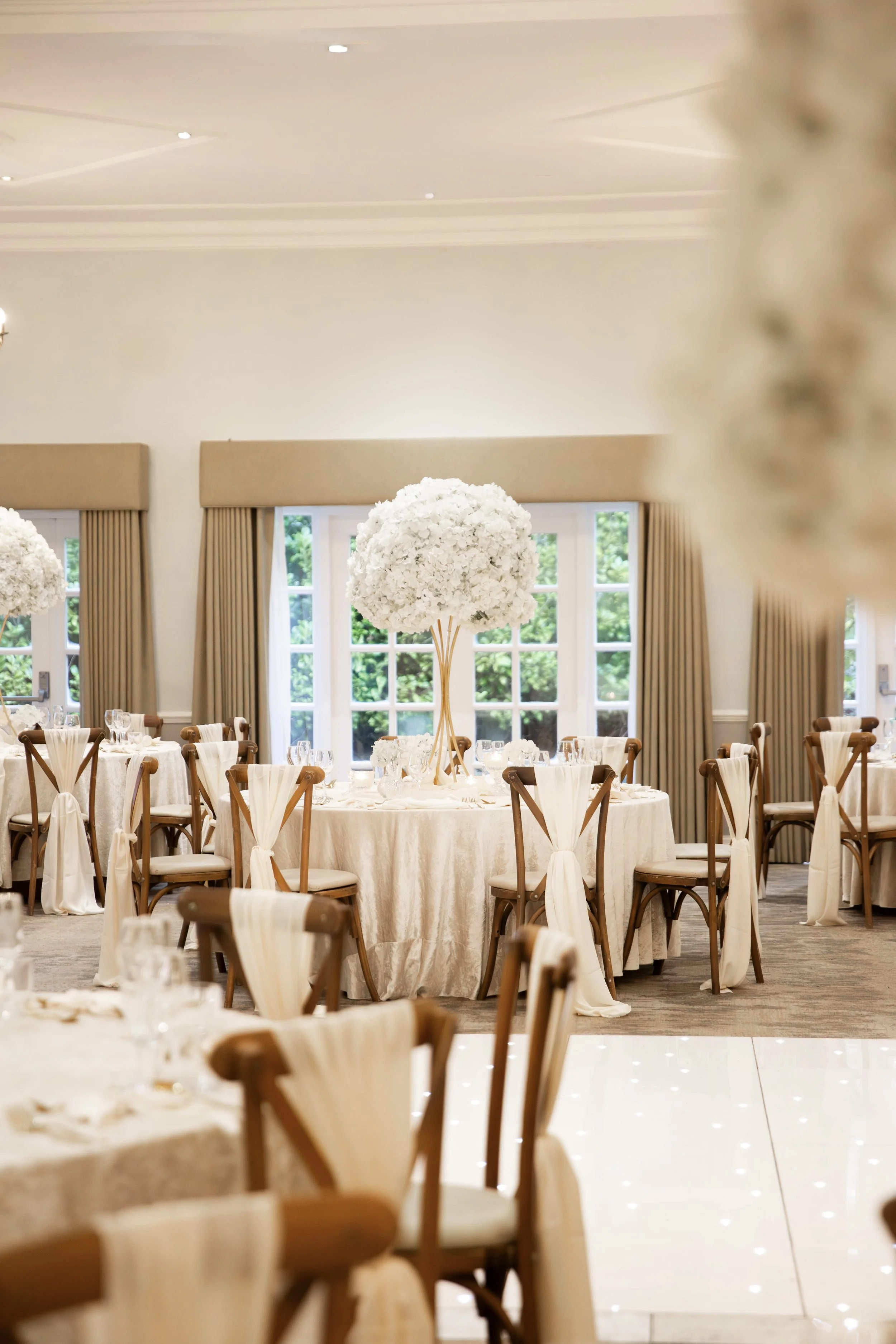 Elegant banquet hall decorated for a formal event with large floral centerpieces on tables, surrounded by chairs with white covers and beige sashes, large windows with beige curtains, and soft lighting.