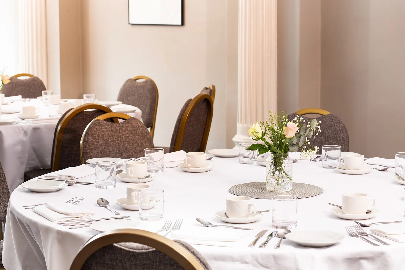 A round banquet table set for a meal with white tablecloth, white cups and saucers, clear glasses, and silverware. A small bouquet of pink and white flowers in a glass jar is in the center of the table, which is on a woven placemat. In the background, additional tables are similarly set, with beige walls and curtains.
