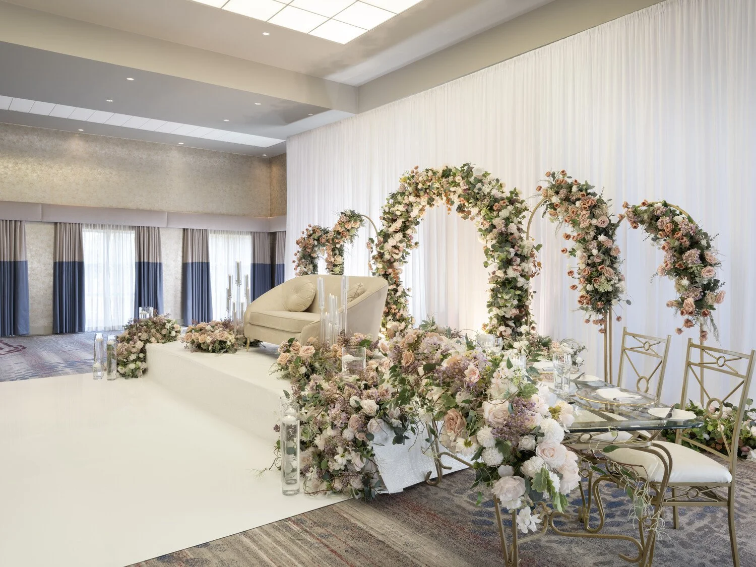 Elegant wedding ceremony setup with a floral arch, a cream loveseat for the bride and groom, and decorated chairs, all adorned with pastel flowers and greenery.