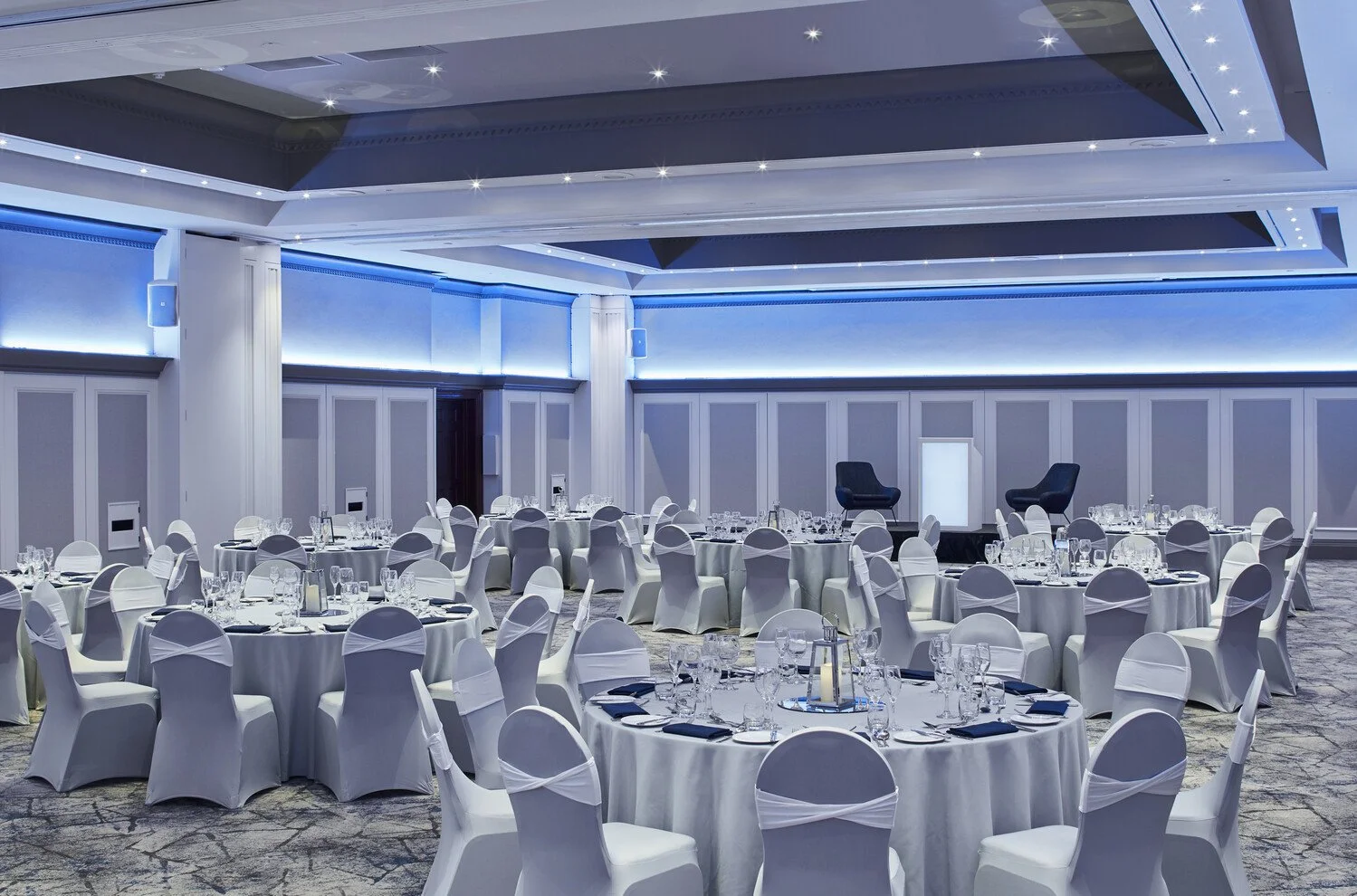 Elegant banquet hall decorated with round tables covered in white tablecloths, set with glassware and black napkins, surrounded by white chairs with white sashes, and featuring a stage with two black chairs in the background, illuminated by blue lighting.