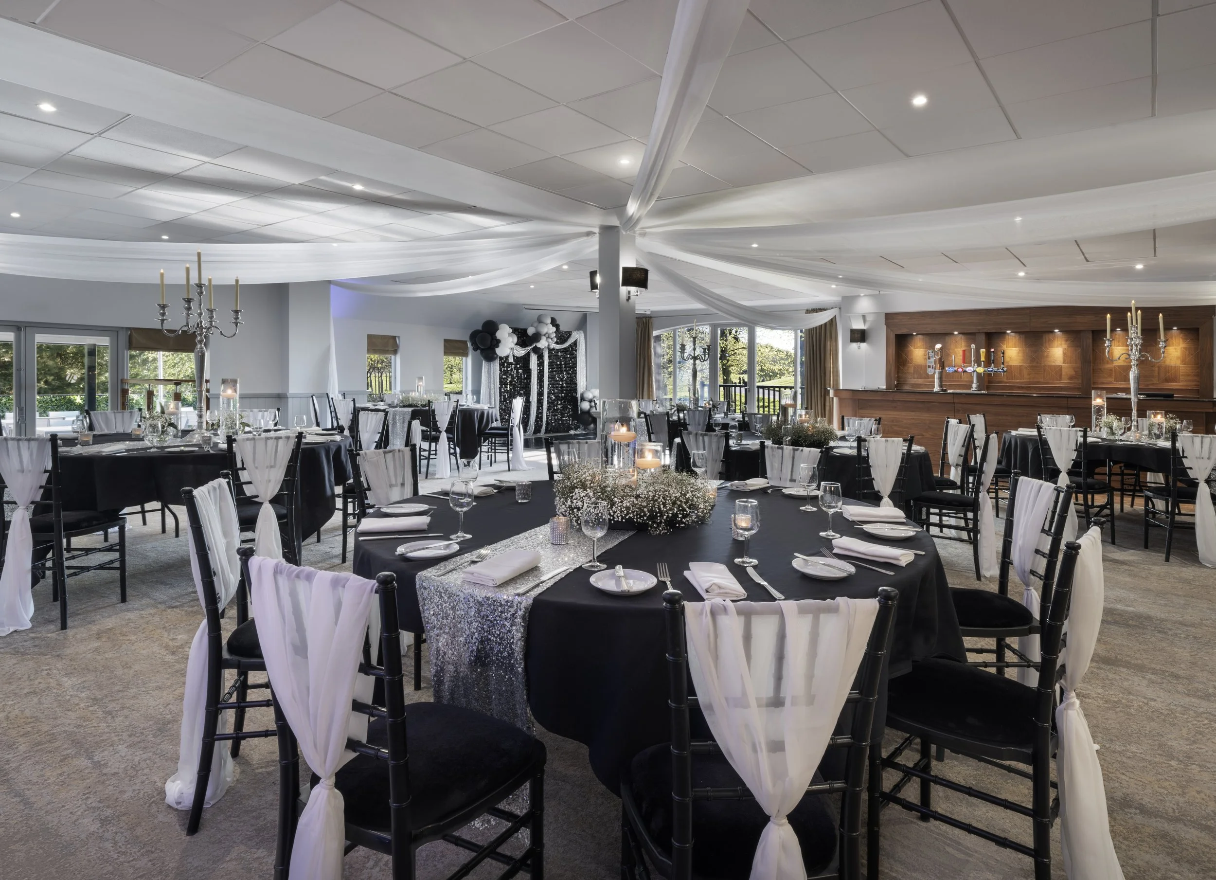 Elegant event hall decorated with white drapery hanging from the ceiling, set up with round tables covered with black and silver tablecloths, adorned with floral centerpieces and candles, large windows with natural light, and a wood-paneled bar area.
