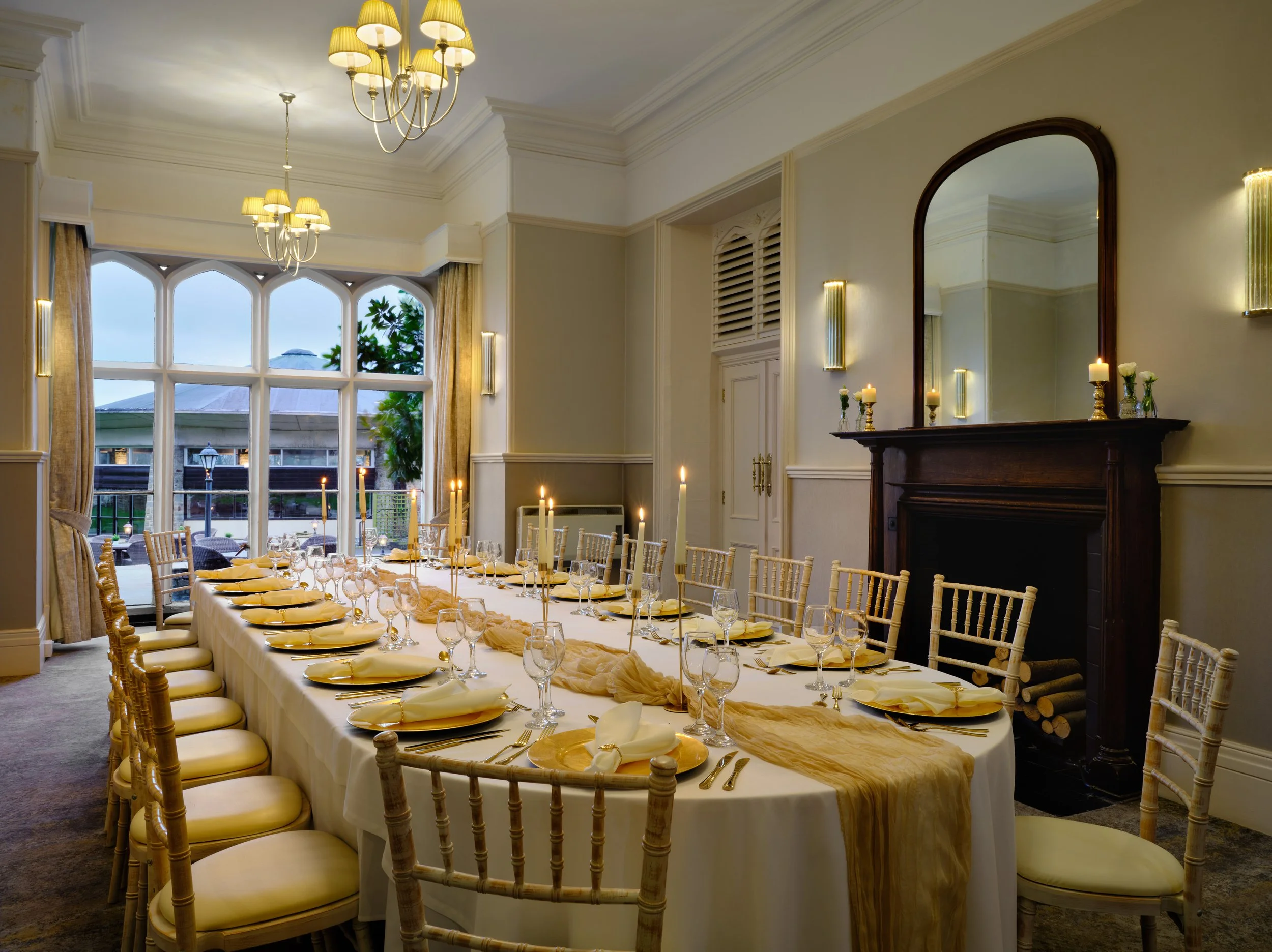 Elegantly decorated dining room with a long table set with yellow napkins, wine glasses, and golden plates. The room has large windows with curtains, chandeliers, and a fireplace with candles and decorative vases, creating a refined atmosphere.