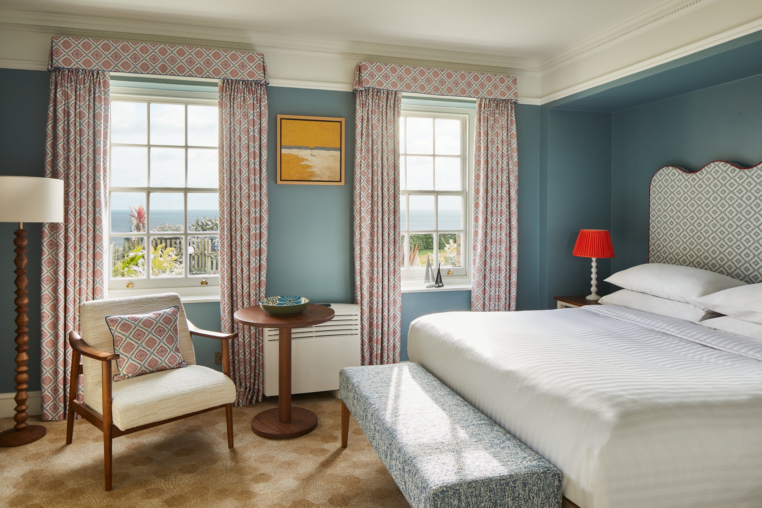 A hotel room with two windows overlooking the ocean, decorated with patterned curtains and a framed seaside picture, featuring a bed with a blue patterned headboard, a red lampshade, and a small sitting area with a chair, table, and decorative pillows.