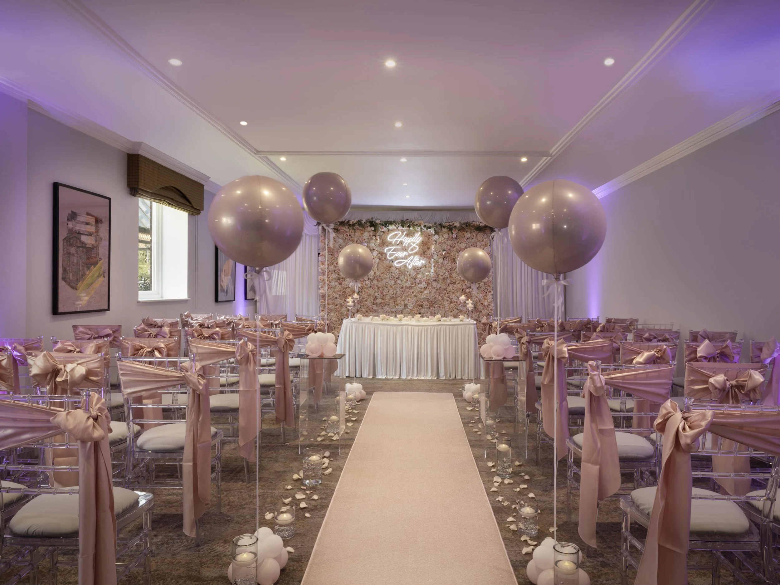 Elegant wedding ceremony setup with chairs decorated with pink satin bows, white flowers, floating candles, a flowered backdrop with a white neon sign that reads 'Happily Ever After', and large pink balloons.