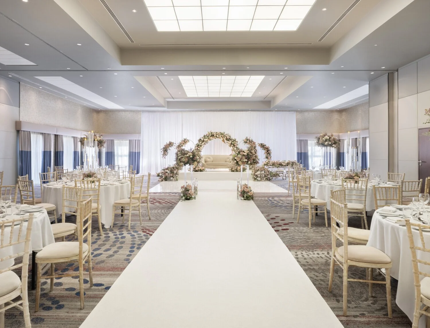 Indoor wedding reception setup with round tables, elegant chairs, and a decorated stage with flowers and a sofa.