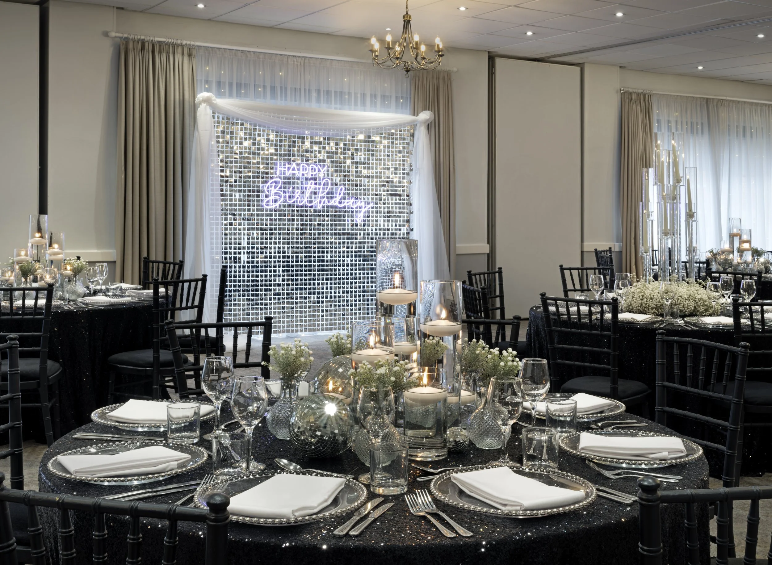Decorated banquet hall with black tables, silver candles, and white floral centerpieces, set for a celebration with a backdrop that says 'Happy Birthday' in neon lights.