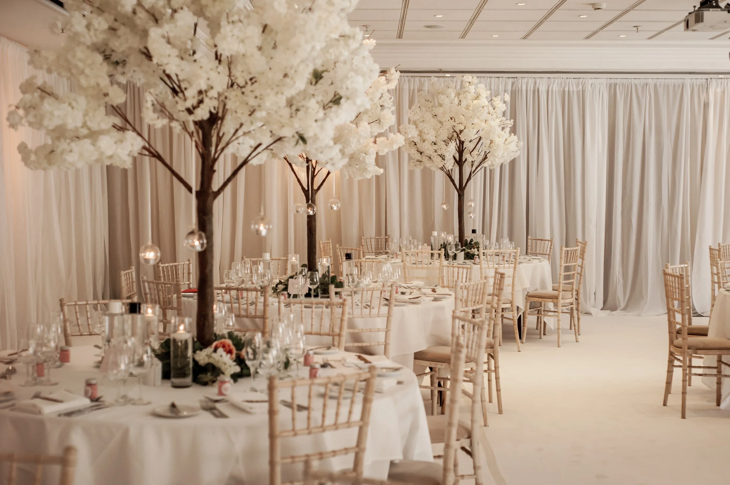 Elegant banquet hall decorated with white flowers on tall trees, round tables set with glassware, candles, and floral centerpieces, surrounded by light wood chairs, with cream-colored drapery and ceiling panels in the background.