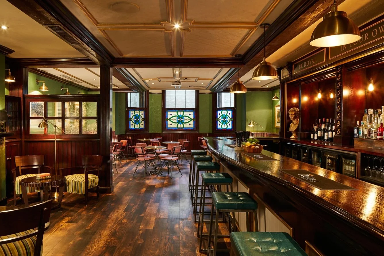 Inside a cozy pub with dark wood furniture, green walls, stained glass windows, and a polished bar counter with bar stools.