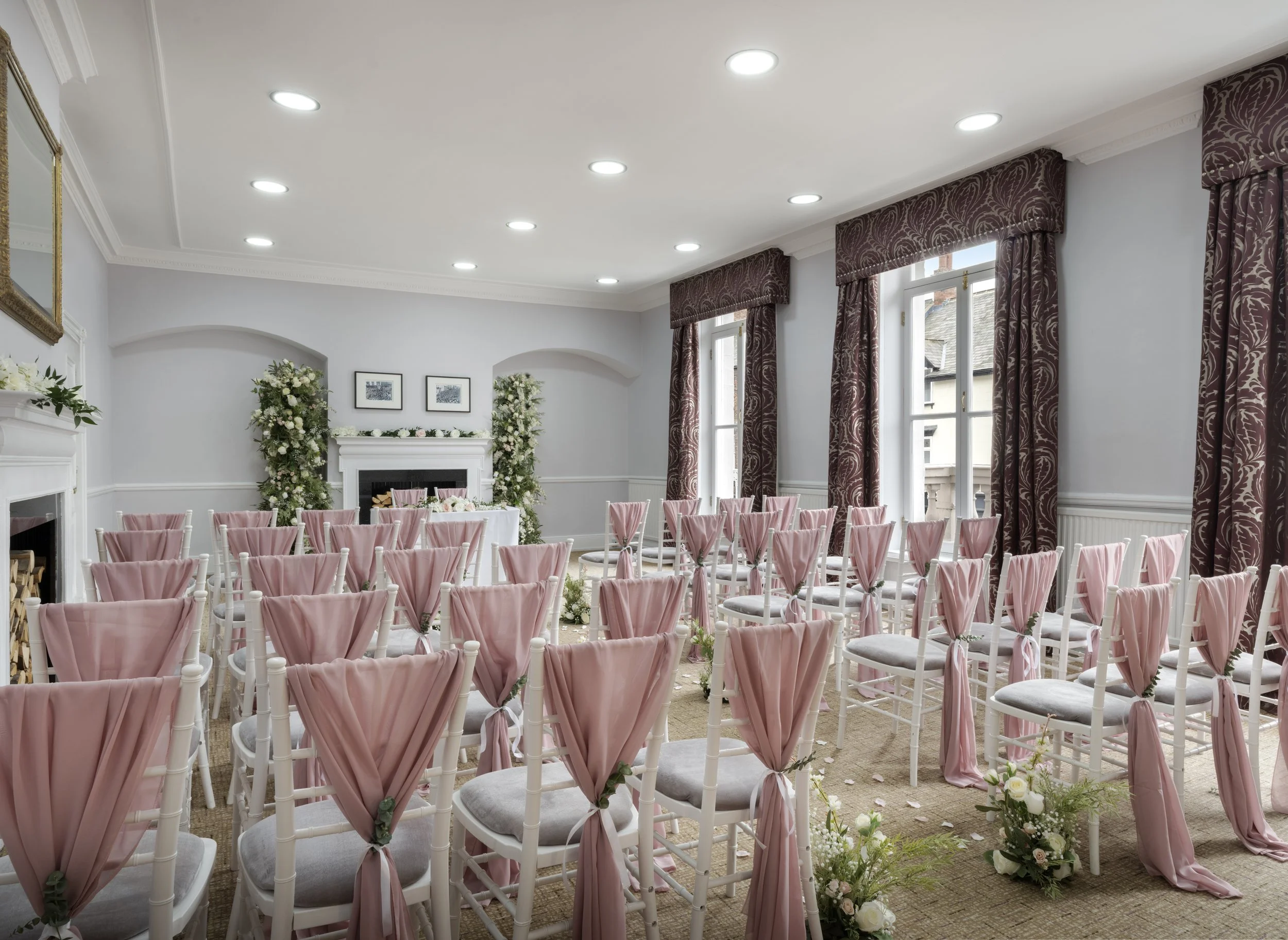 An elegant indoor wedding ceremony setup with rows of chairs draped with pink fabric, facing a decorated fireplace with floral arrangements, and large windows with purple-patterned curtains.