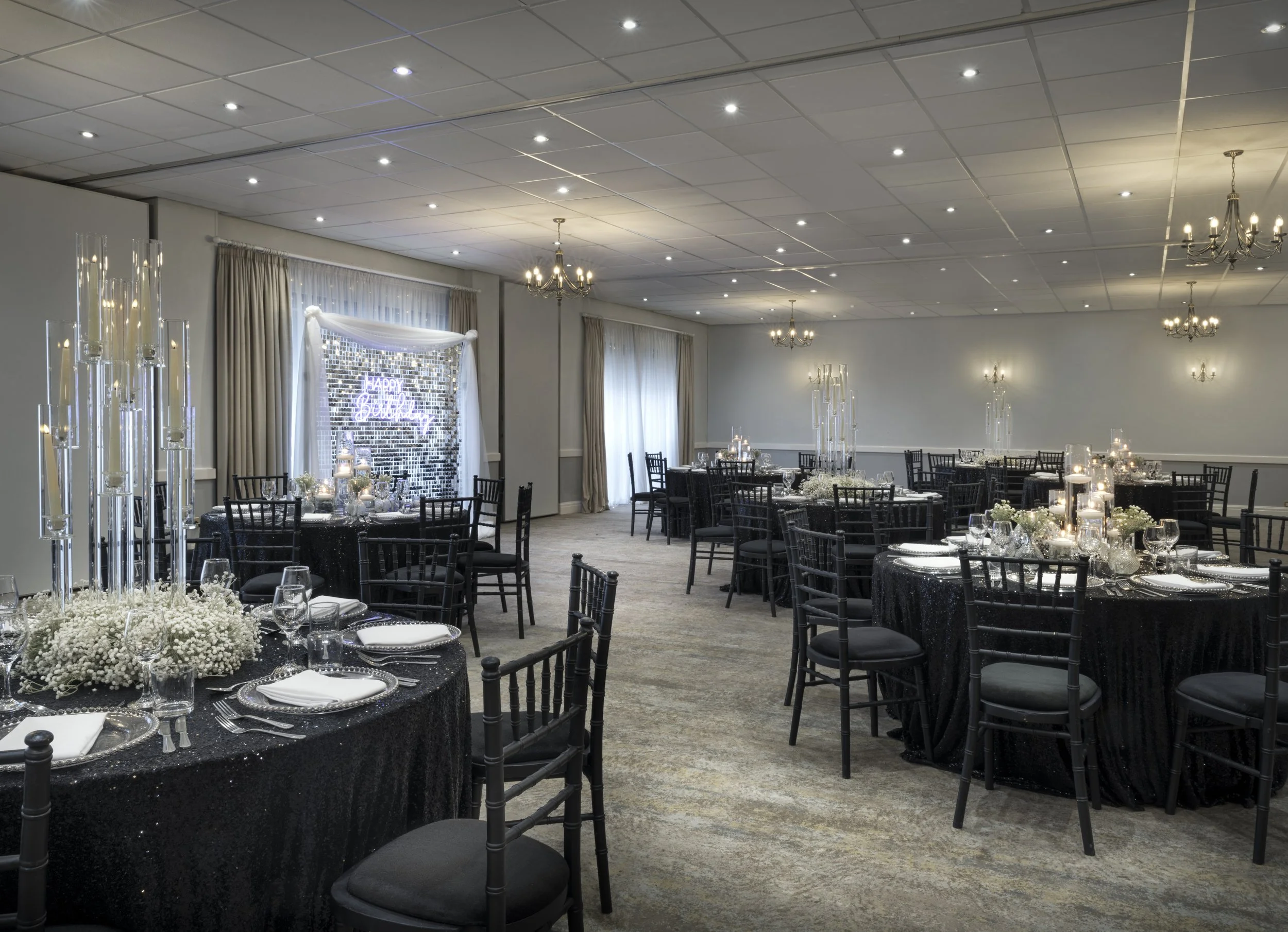 Event hall decorated with tables, flowers, and candles for a celebration or wedding, with a neon sign that says 'Happy Birthday' on a brick wall in the background.