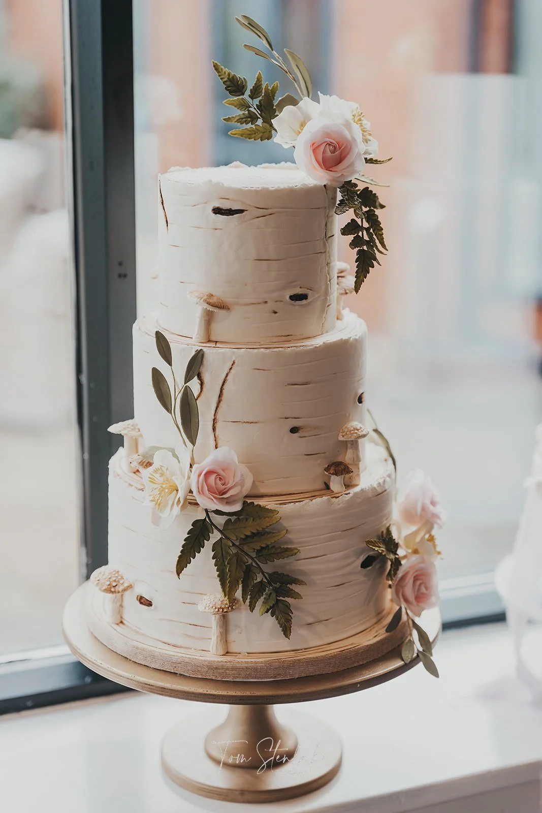 A three-tiered rustic wedding cake decorated with pink roses, green leaves, and small mushrooms, placed on a wooden cake stand near a window.