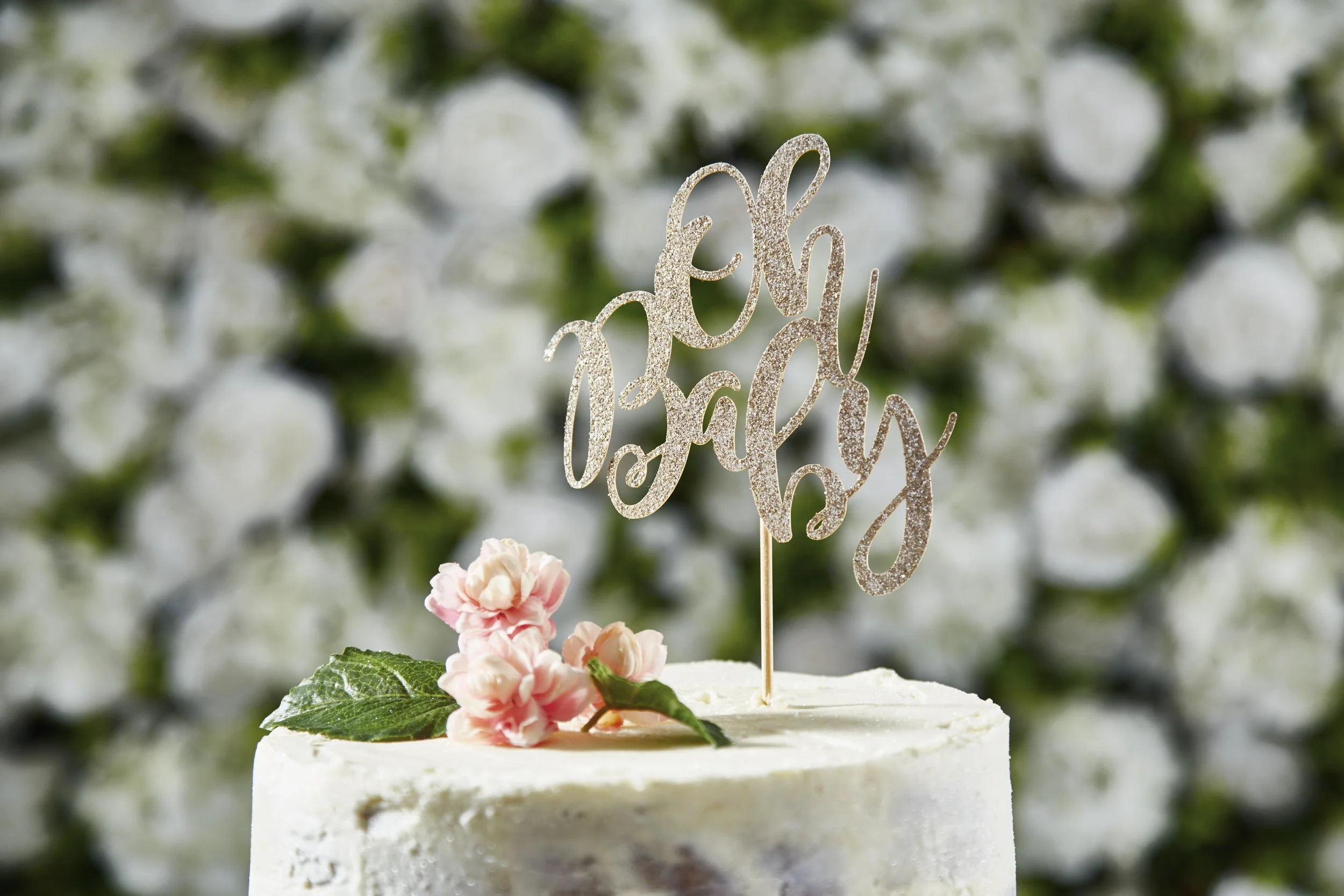 A white cake decorated with pink flowers and green leaves, with a glittery gold cake topper that says "oh happy day". The background consists of out-of-focus white flowers and greenery.