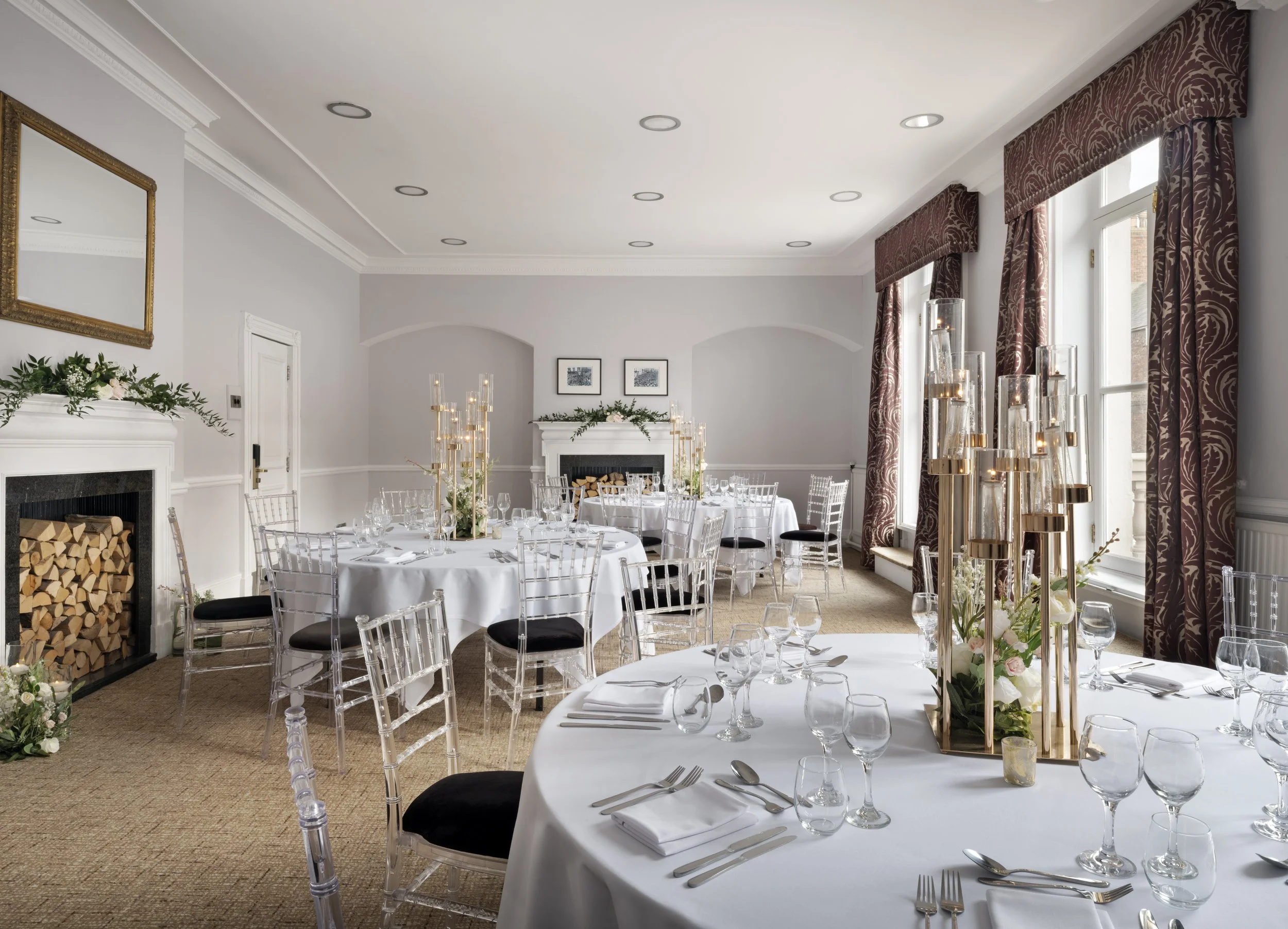 Elegant banquet room decorated for a formal event with white tablecloths, glassware, floral centerpieces, and transparent chairs, featuring tall candle holders, a fireplace with logs, framed artwork, ornate curtains, and a ceiling with recessed lighting.