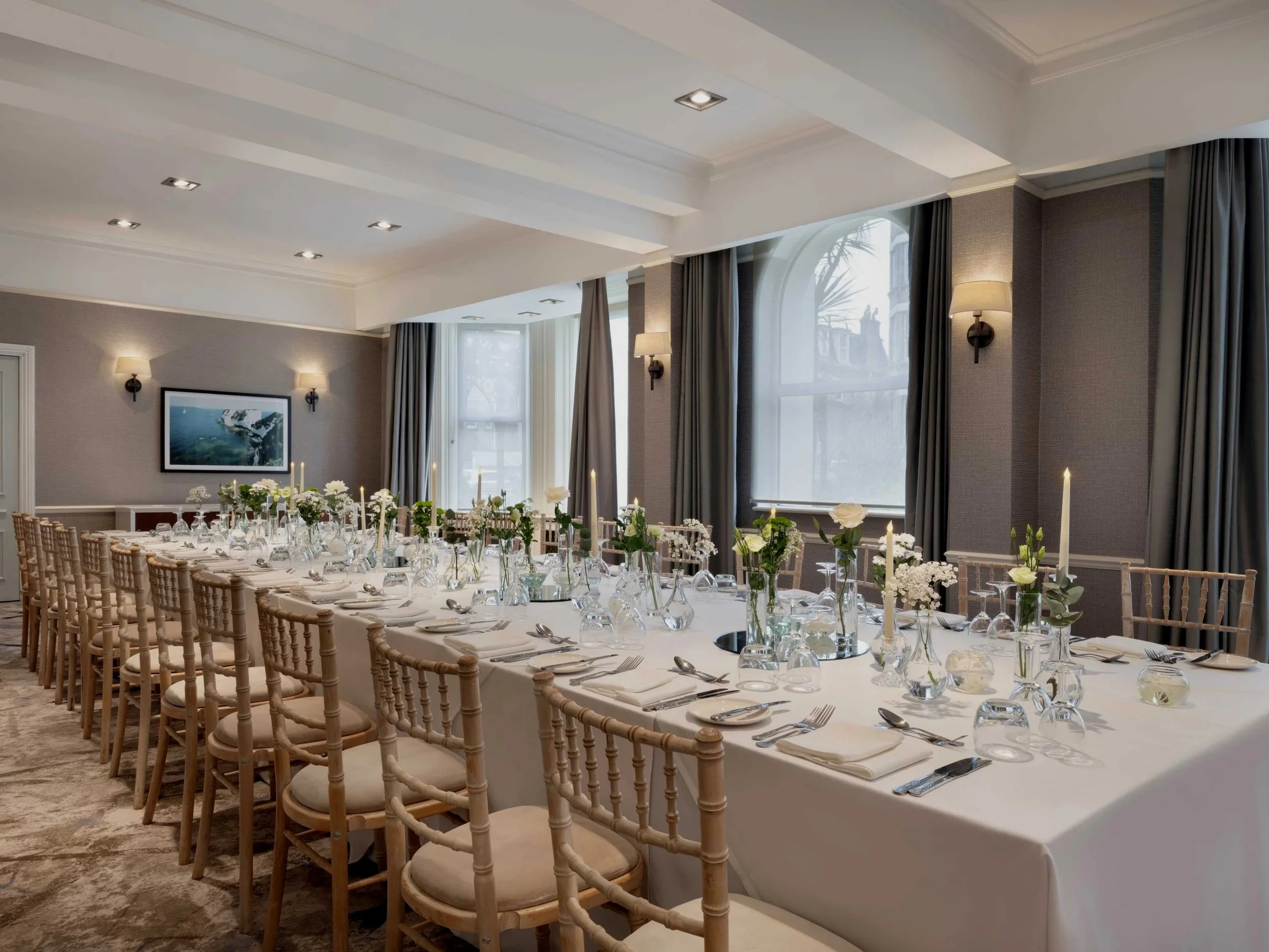 Elegant banquet room set for a wedding or special event, with long table, white tablecloth, candle centerpieces, flower arrangements, and wooden chairs, illuminated by wall sconces and natural light from large windows.