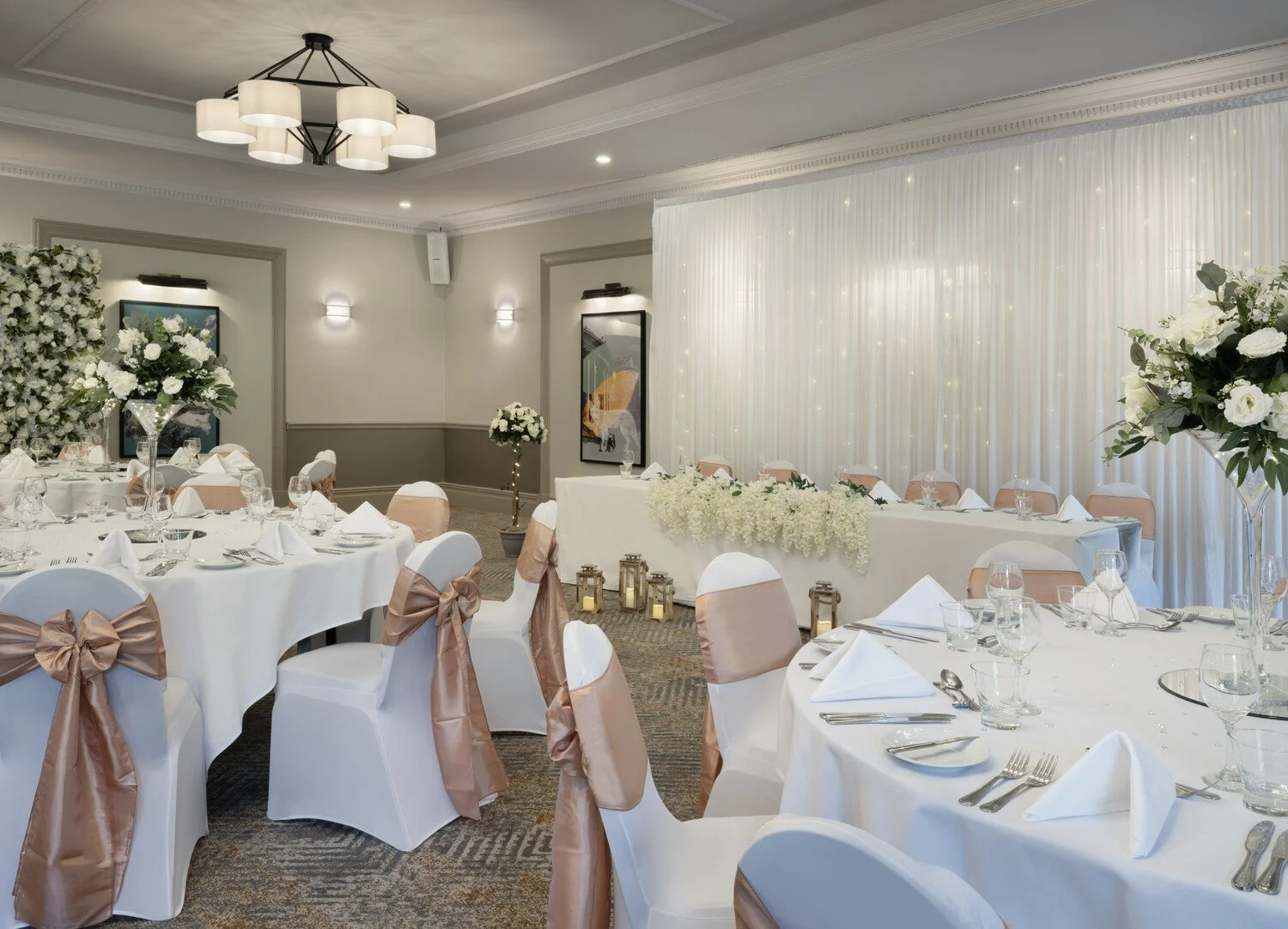Elegantly decorated banquet hall for a wedding reception with white tables, peach satin bows on the chairs, floral centerpieces, white curtains with string lights, and a decorated head table.
