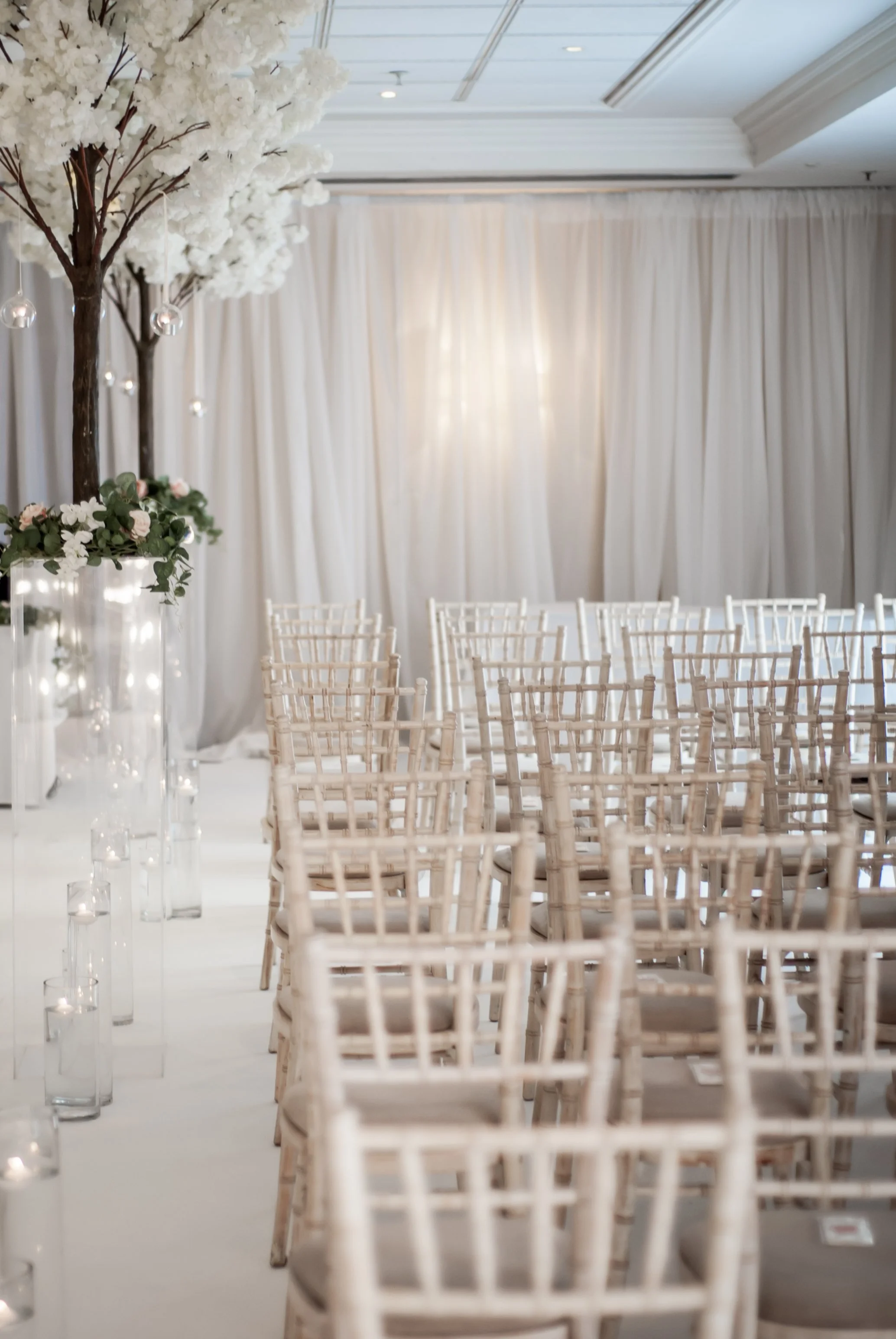 Wedding ceremony setup with rows of white chairs and tall floral centerpieces with white flowers and greenery, decorated with hanging glass orbs, and white drapery backdrop in an indoor venue.