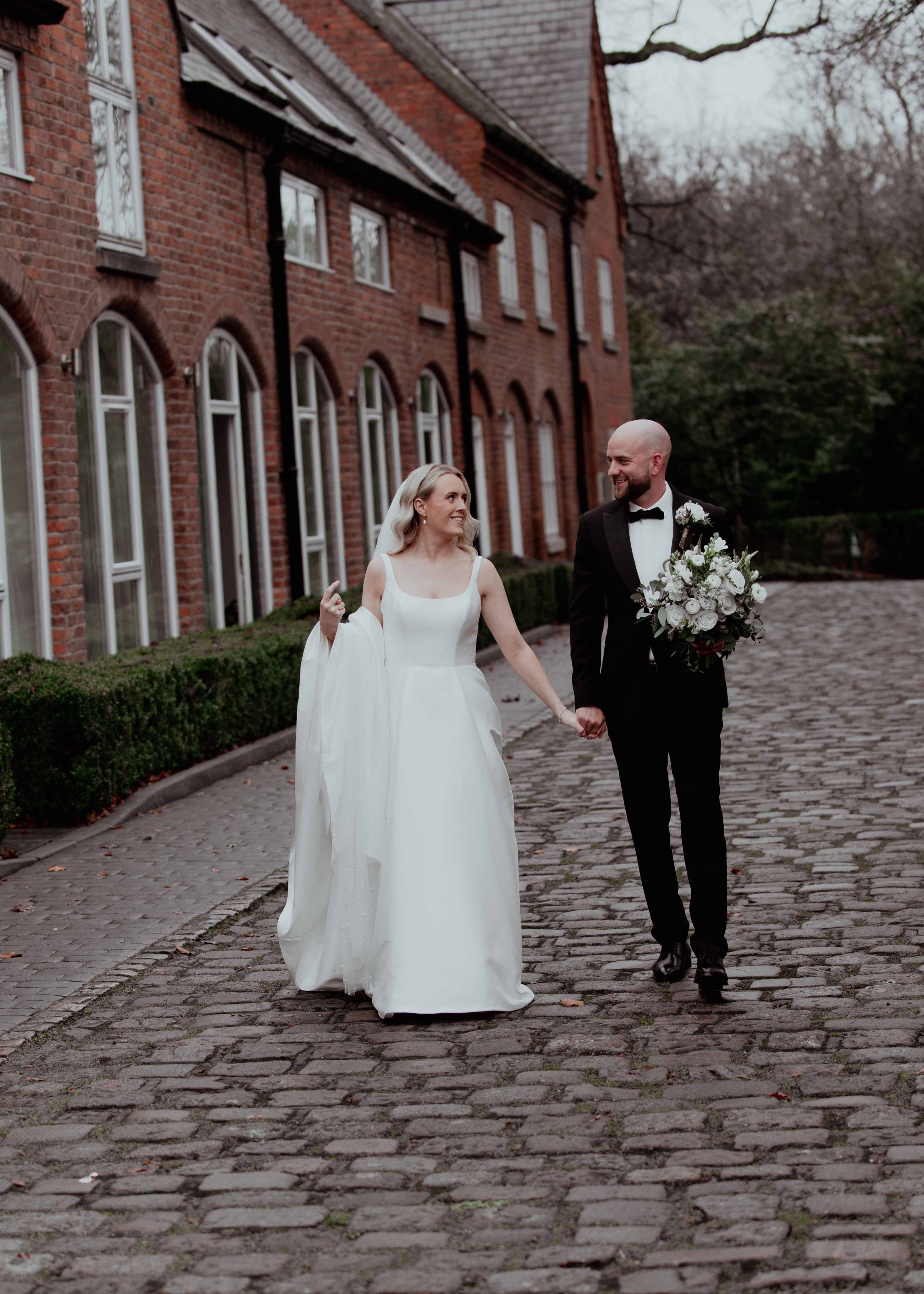 A bride and groom walking hand in hand outdoors on a cobblestone path beside a red brick building, with trees in the background. The bride wears a white wedding dress with a veil, and the groom wears a black tuxedo with a bow tie and holds a bouquet of white flowers.